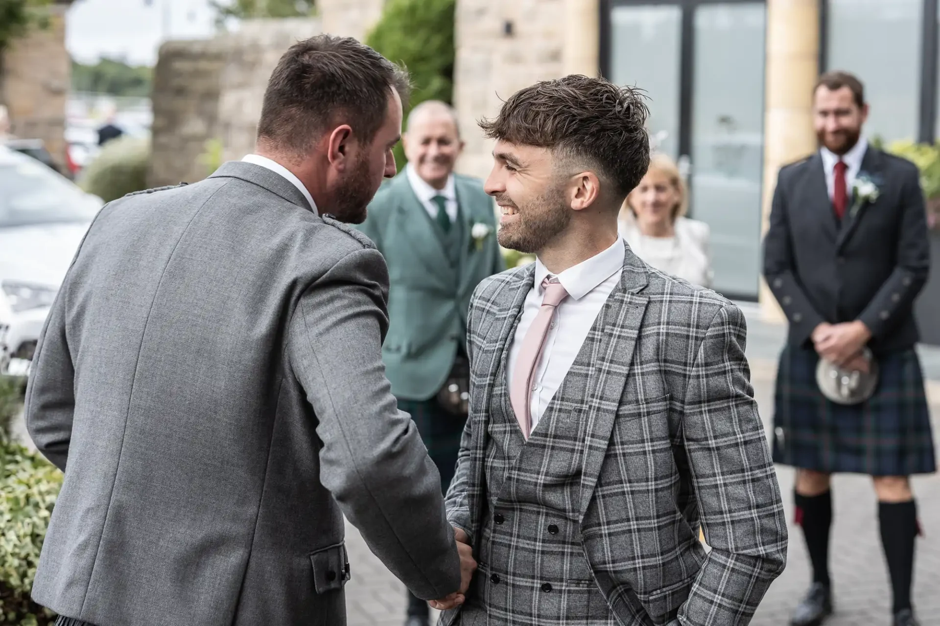 Two men in gray suits shake hands outside, with three people watching in the background.