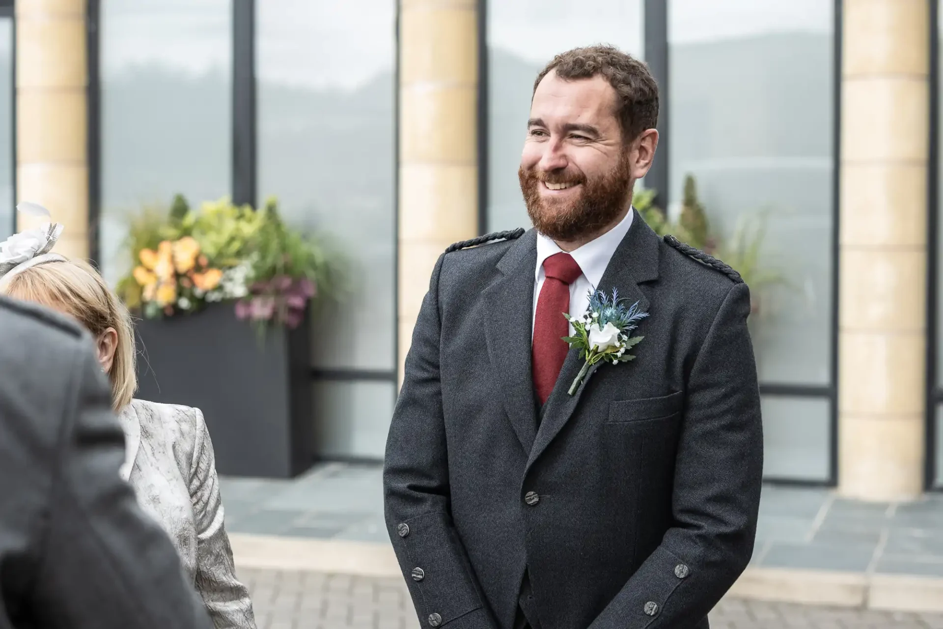 A man in a suit with a boutonniere smiles outside near large windows and potted plants.