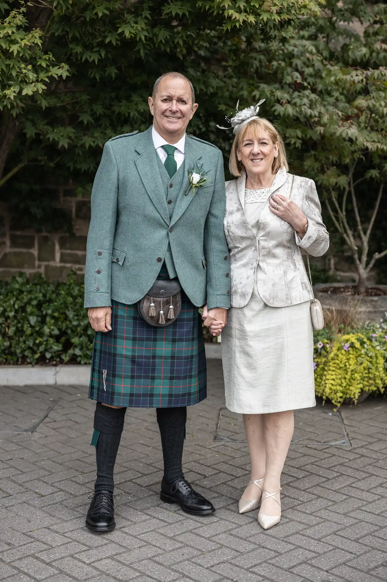 A couple stands outdoors, the man wearing a kilt and green jacket, and the woman in a light-colored jacket and skirt suit. They hold hands and smile at the camera.