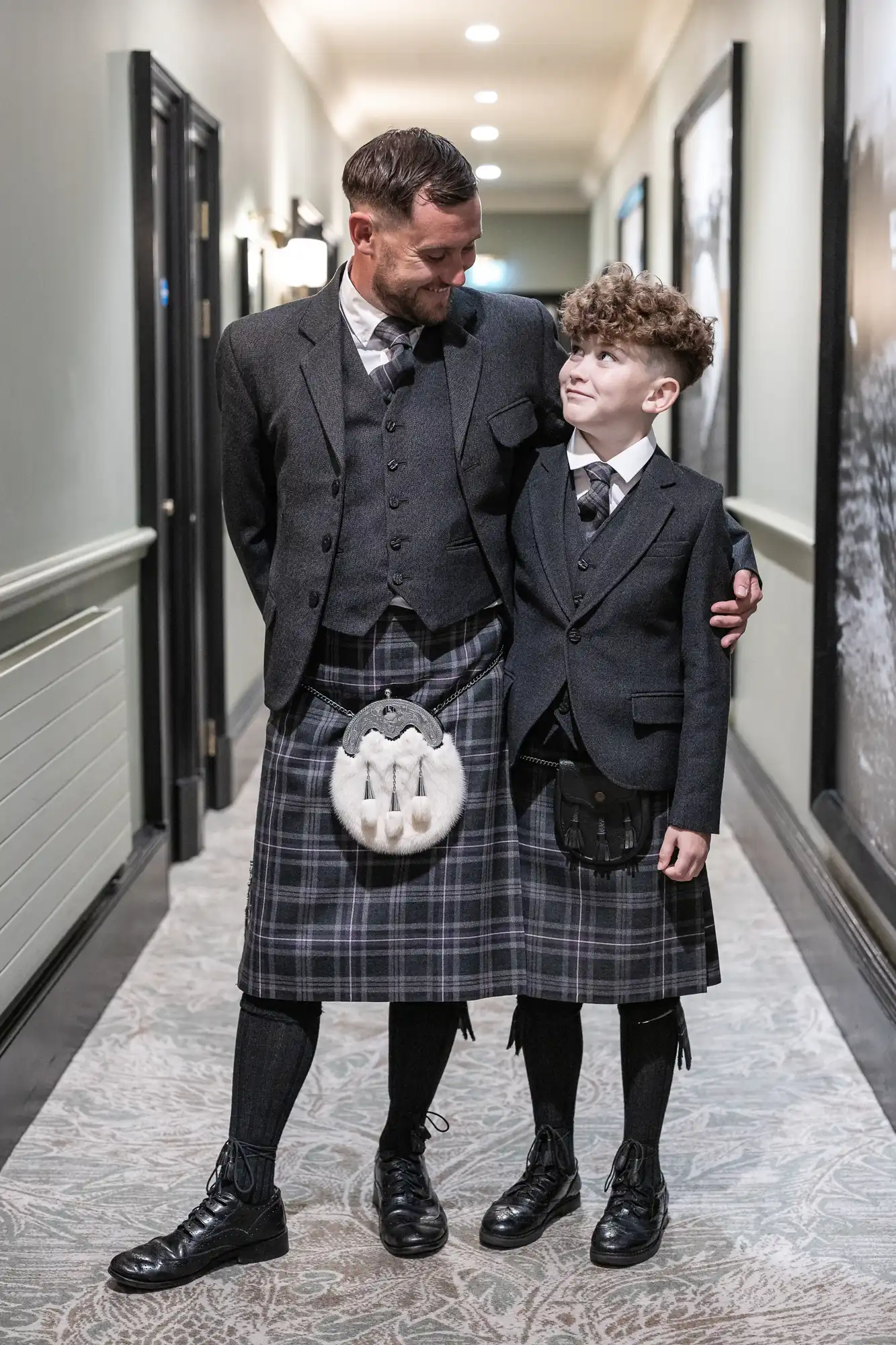 Two people, possibly a father and son, standing in a hallway. Both wear matching formal Scottish attire with kilts and jackets, looking at each other and smiling.