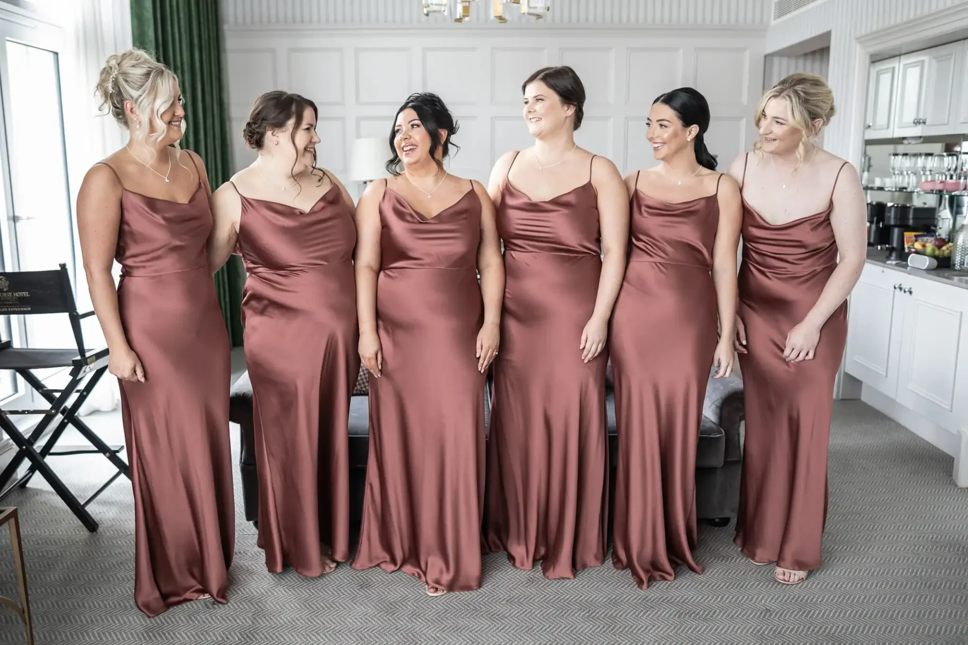 Six women in matching satin dresses stand indoors, smiling and facing each other.