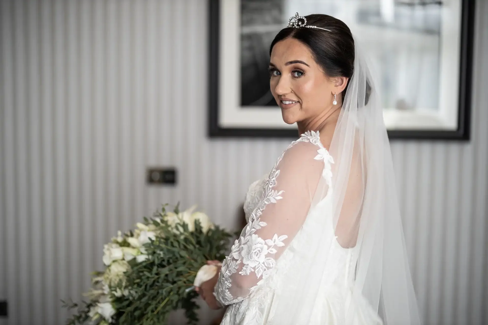 Bride in a white dress with lace sleeves and a veil, holding a bouquet of white flowers, looking over her shoulder in a room with a striped wall and framed picture.