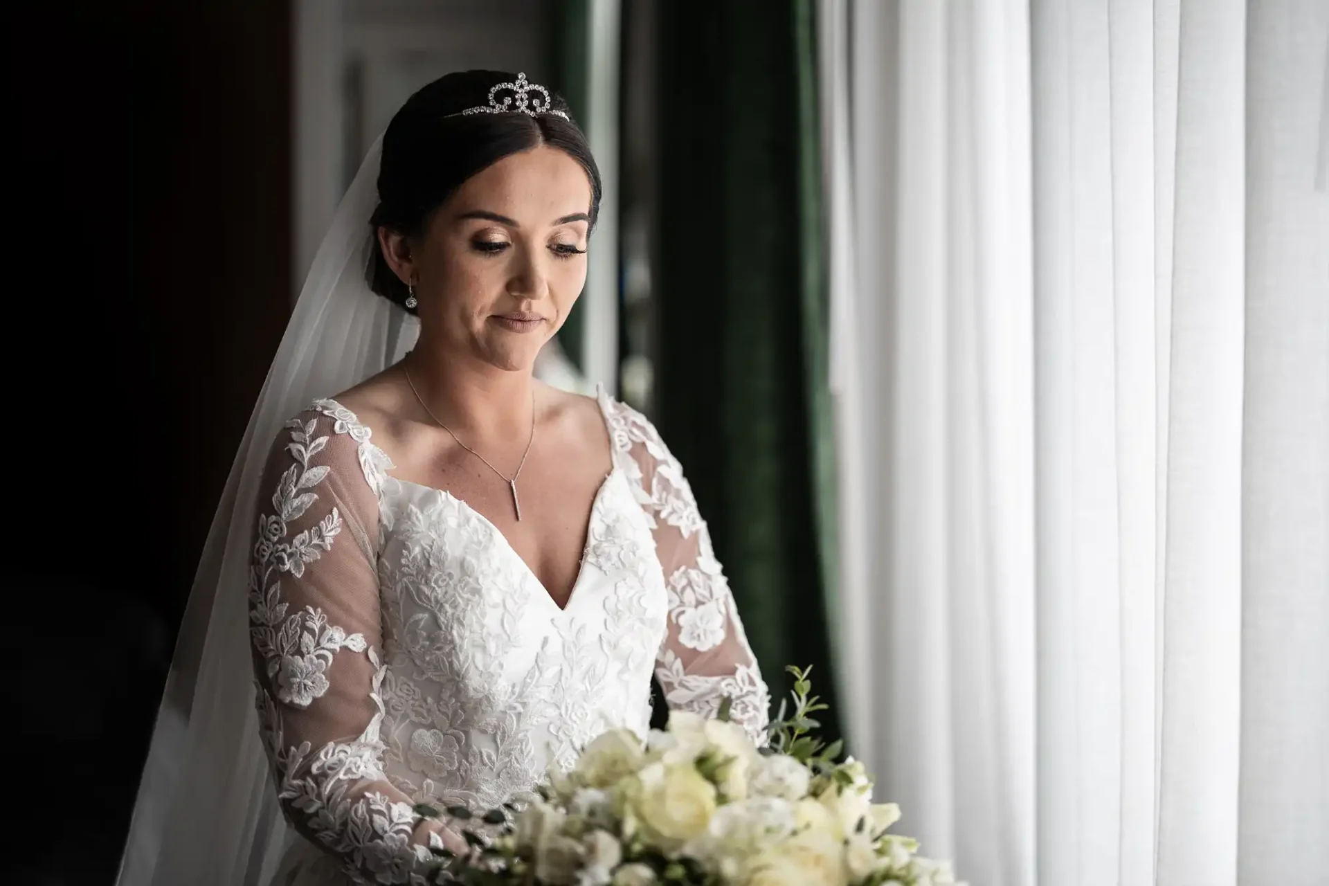 A bride in a white lace wedding dress and veil holds a bouquet of white flowers, standing by a window with sheer curtains.