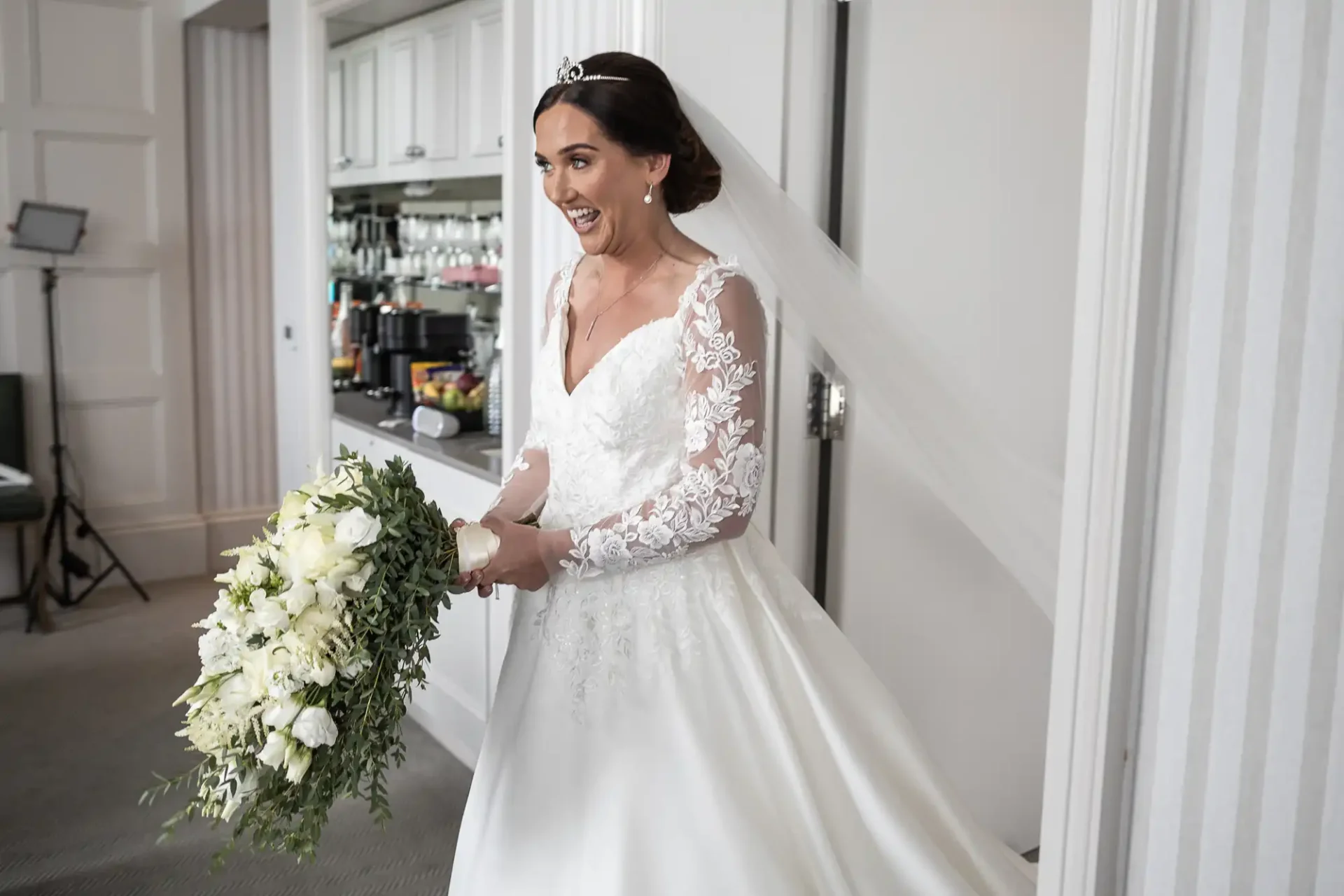 Bride in white gown with lace sleeves and long veil holds a bouquet of white flowers, smiling brightly in a room with white walls and cabinets.