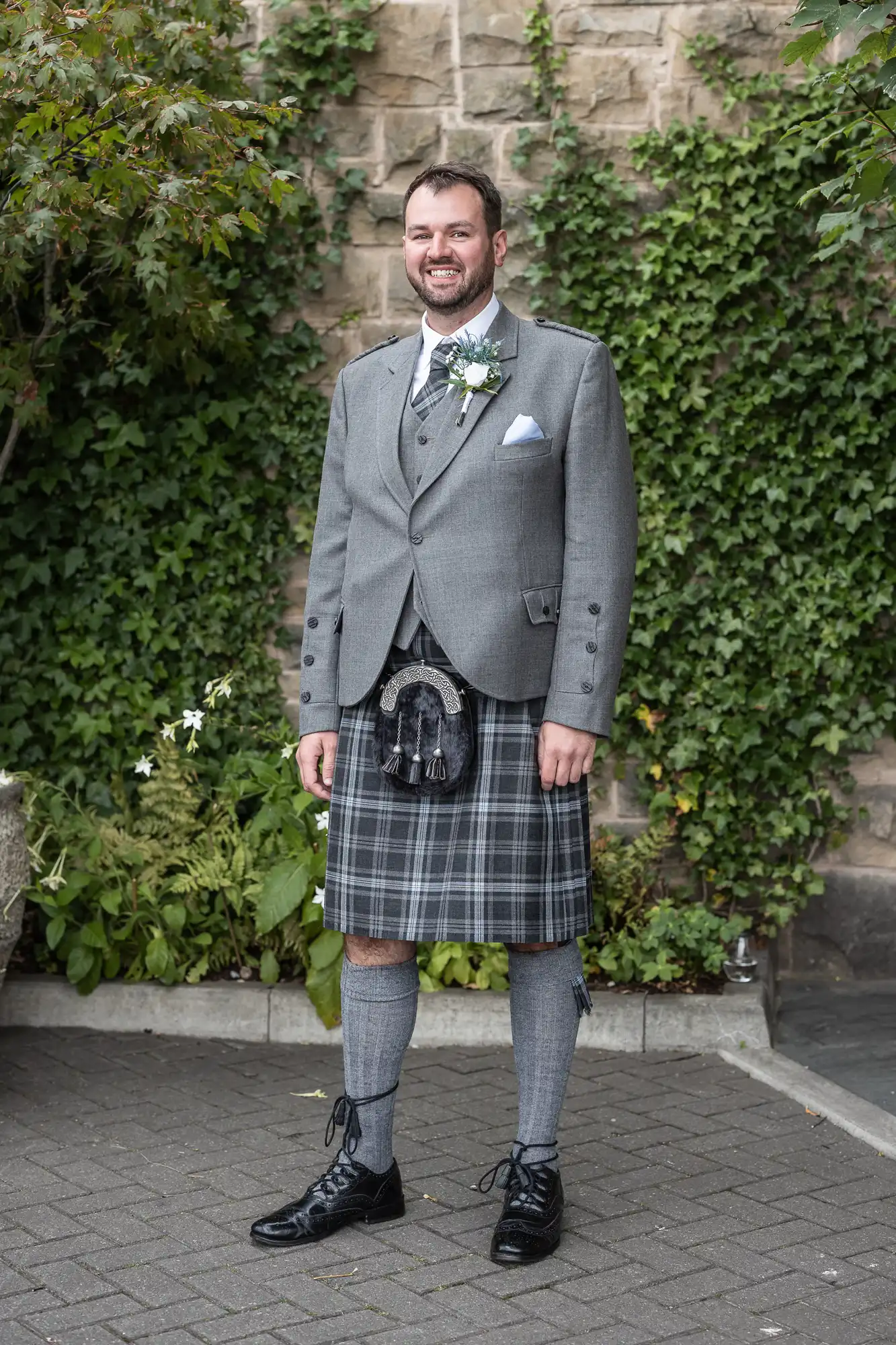 Man wearing a gray suit jacket, white shirt, and tartan kilt stands on a stone pavement with greenery in the background.