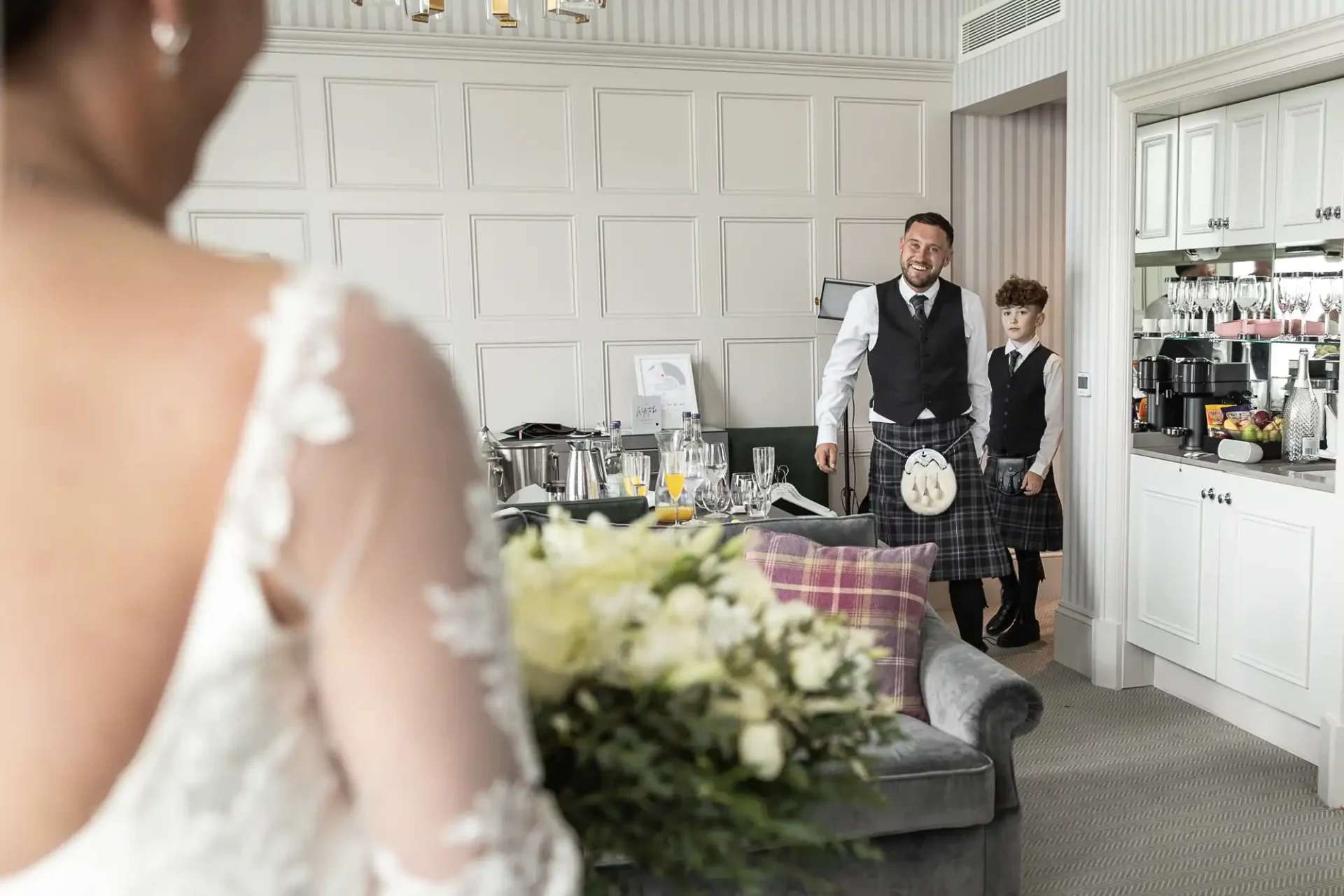 Bride with bouquet facing a man and a boy dressed formally. The room is elegantly decorated with glasses and framed photos on a cabinet.