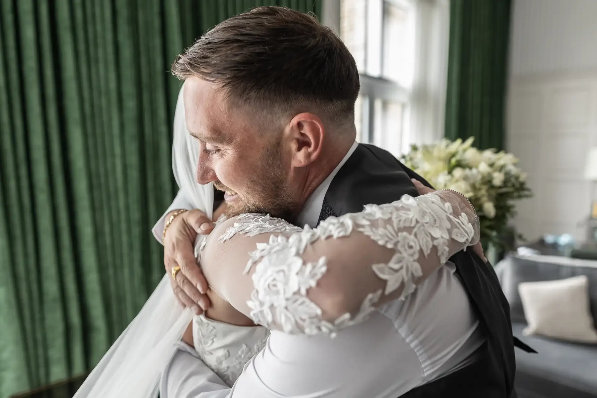 A groom in a suit hugs a bride in a white lace gown indoors with green curtains and a bouquet in the background.