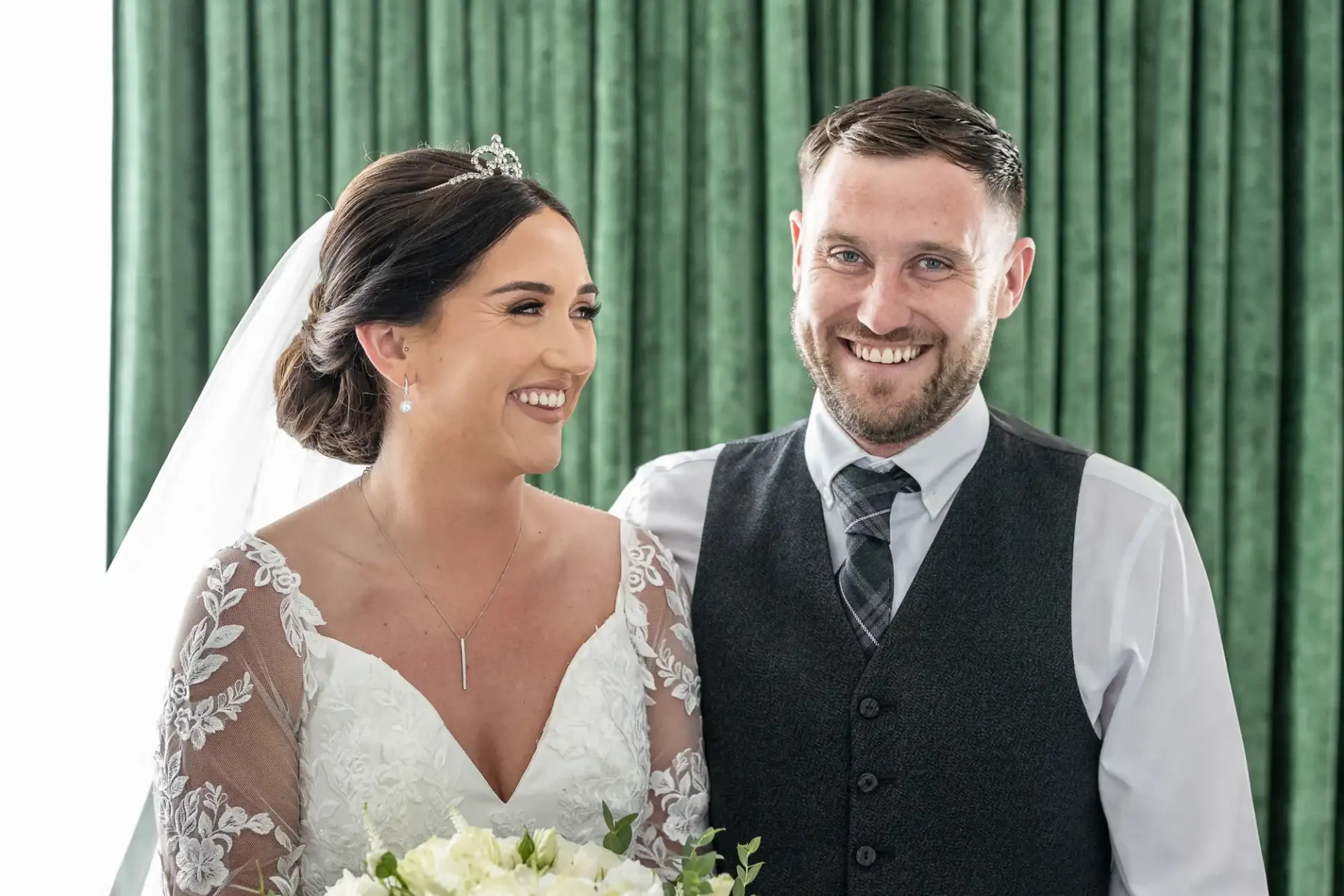 A bride and groom smile, standing together in front of green curtains. The bride holds a bouquet and wears a veil and tiara; the groom is in a vest and tie.