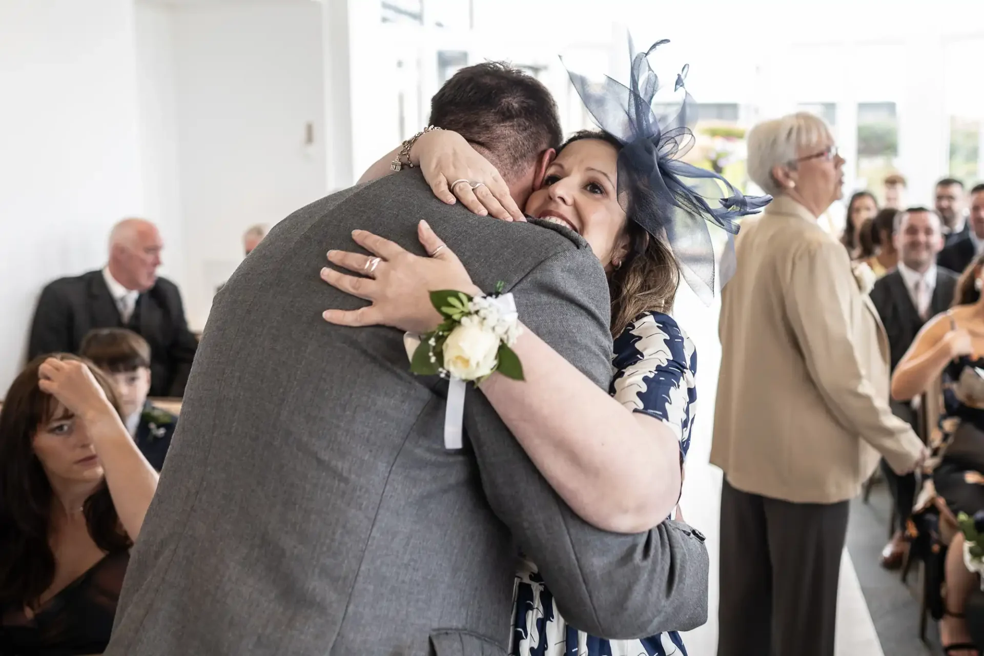 A man in a gray suit and a woman in a patterned dress with a large fascinator hug in a bright room. Onlookers are seated in the background.