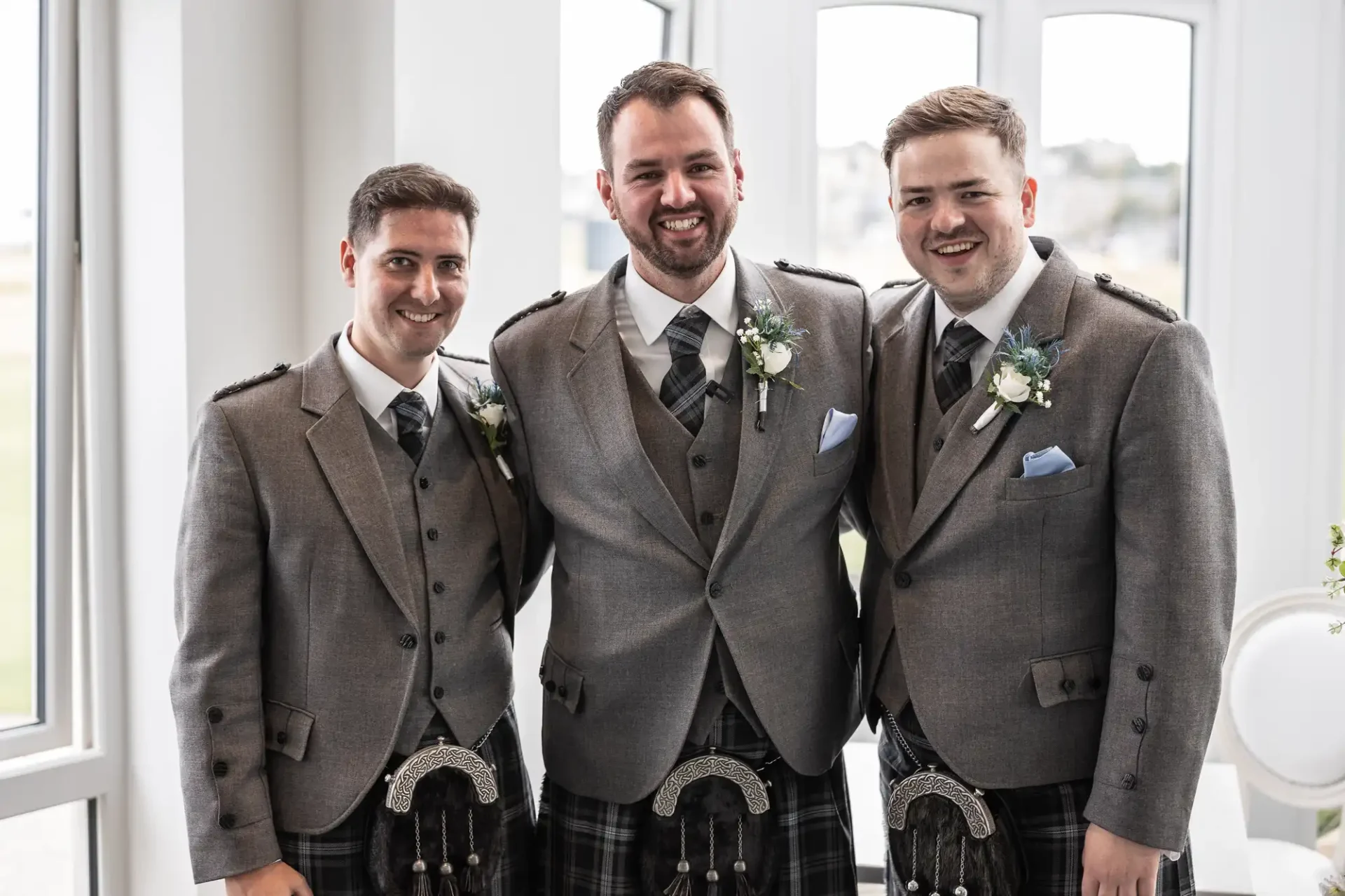 Three men wearing grey suits and traditional Scottish kilts stand together smiling in a bright room.