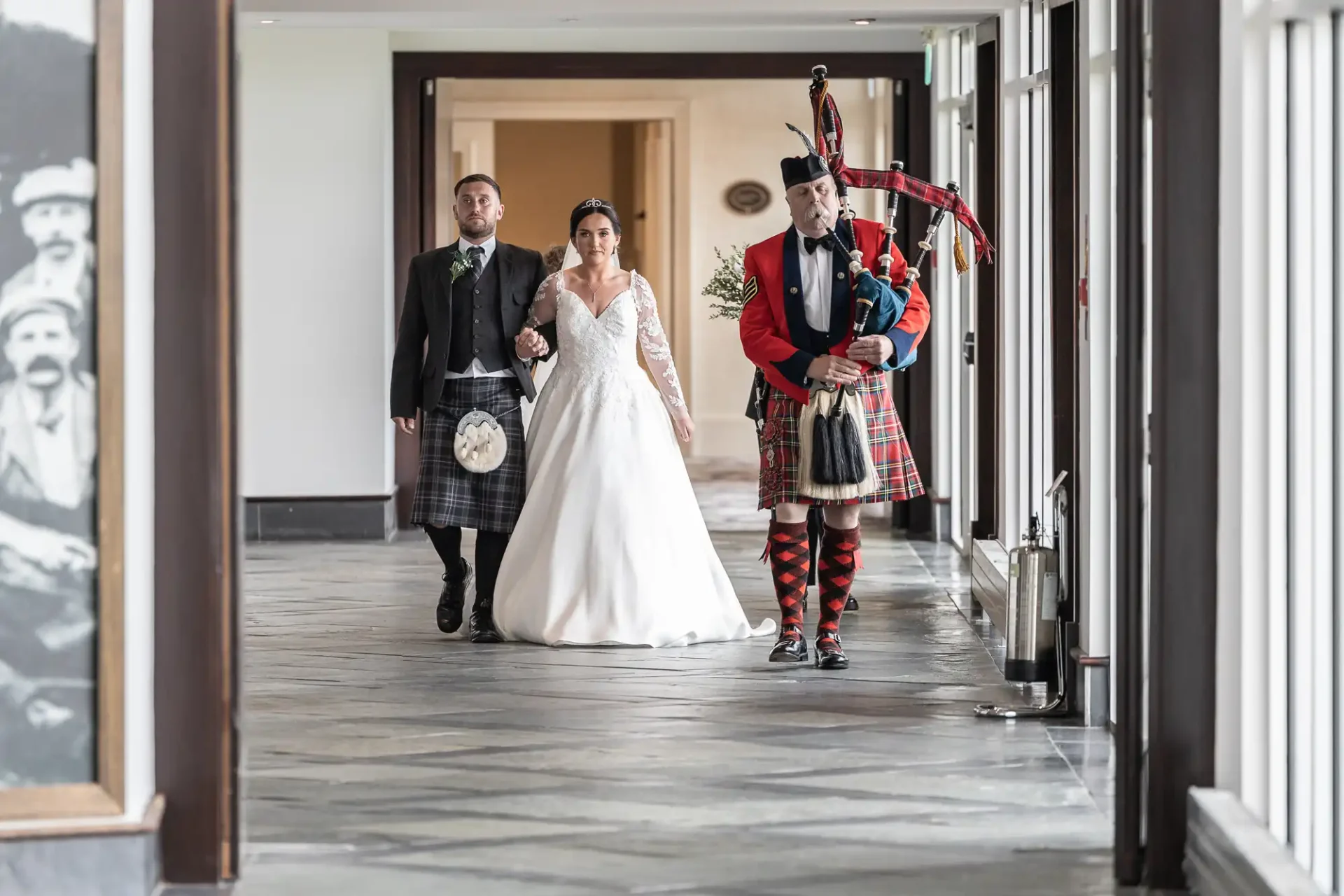 Bride in white gown and groom in Scottish attire walk down a corridor led by a bagpiper in traditional Scottish dress.