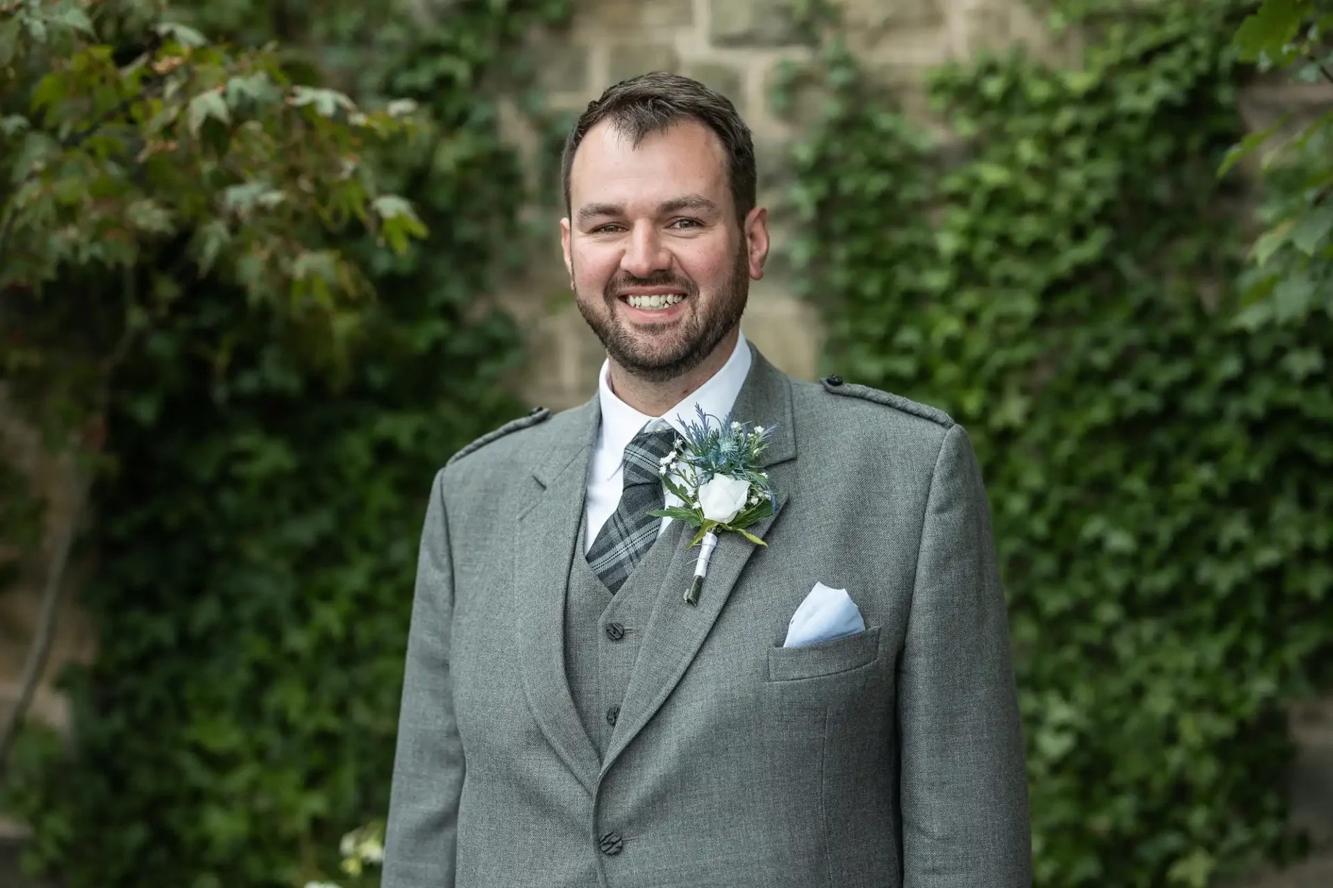 A man in a gray suit with a patterned tie and boutonniere smiles, standing in front of an ivy-covered stone wall.