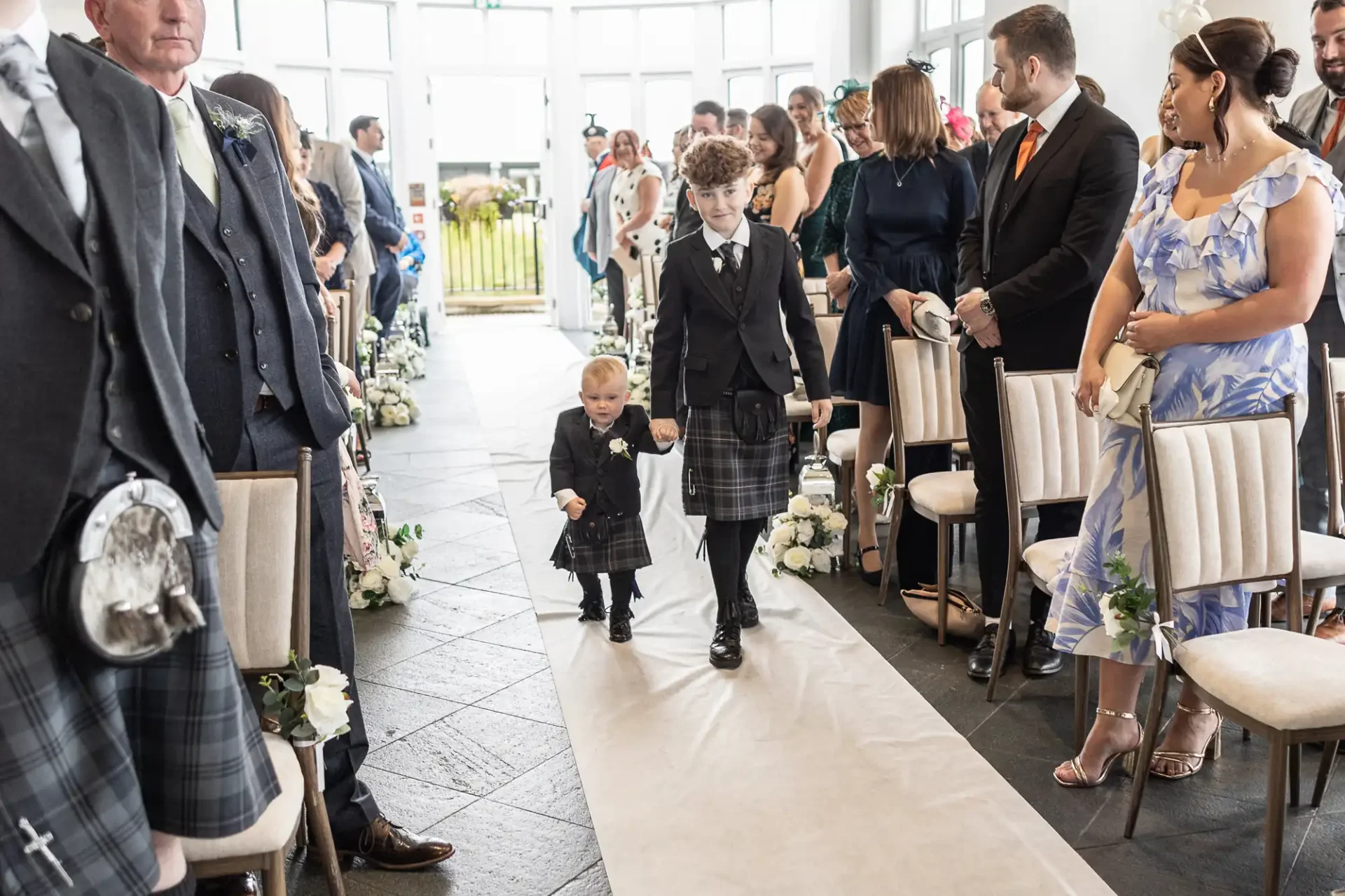 Two children in kilts walk down an aisle in a wedding ceremony, surrounded by seated guests.