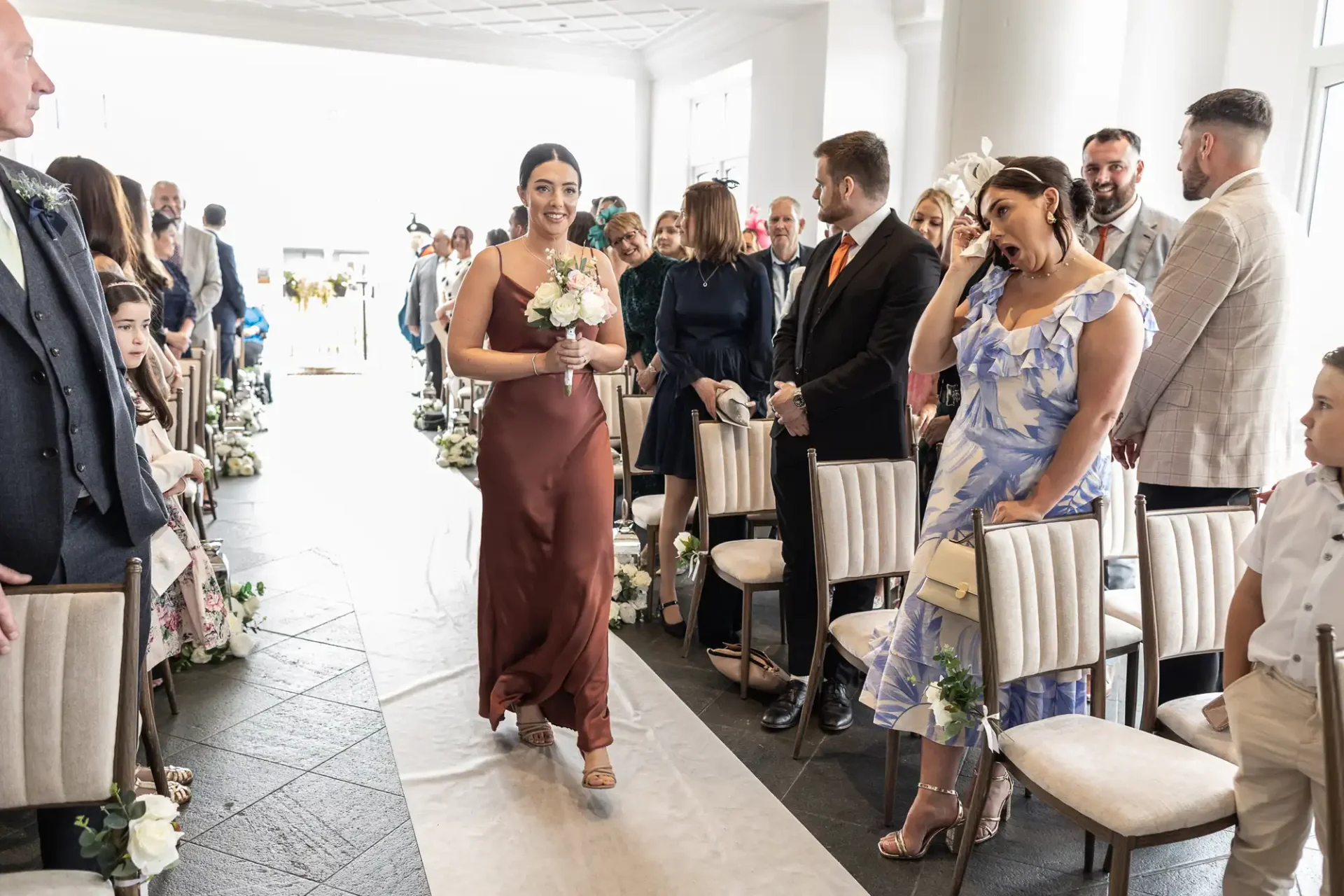 A woman in a brown dress walks down the aisle holding flowers at a wedding, with seated guests on either side.