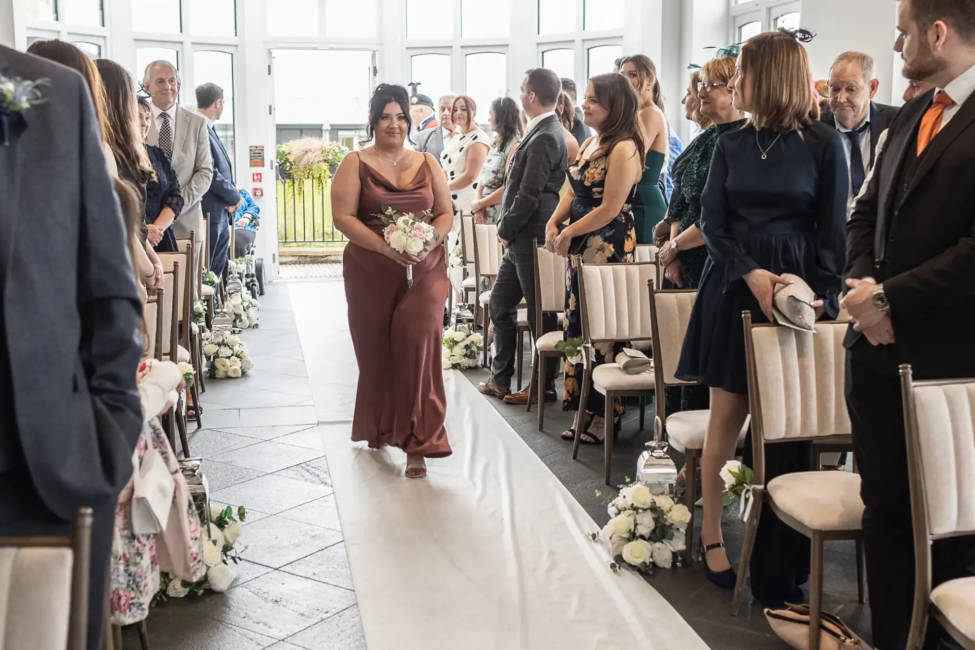 A woman in a brown dress walks down an aisle at a wedding ceremony, holding a bouquet. Guests seated on either side observe.