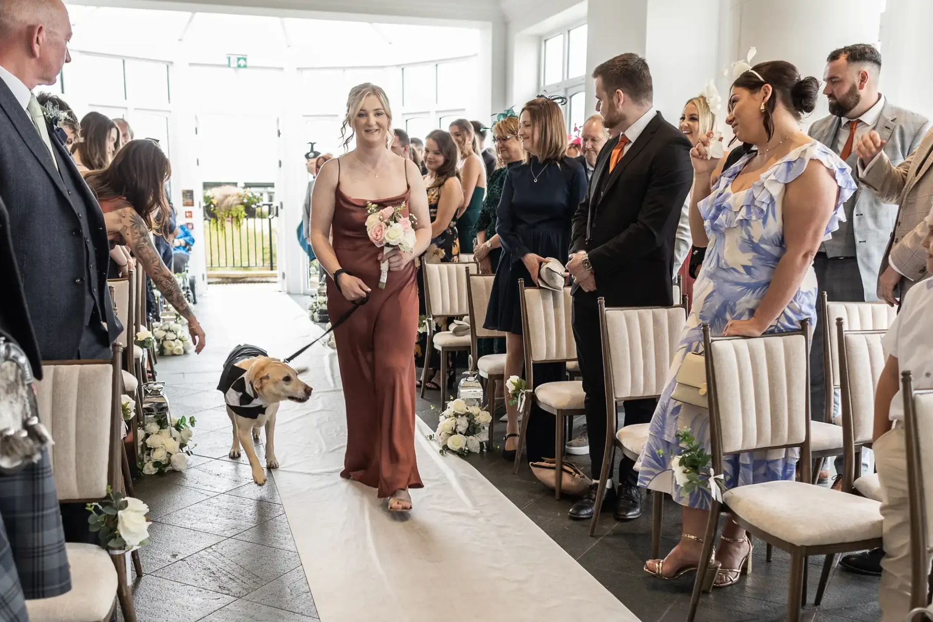 A woman in a brown dress walks down the aisle holding flowers, accompanied by a dog in a suit. Guests seated on either side observe the procession.