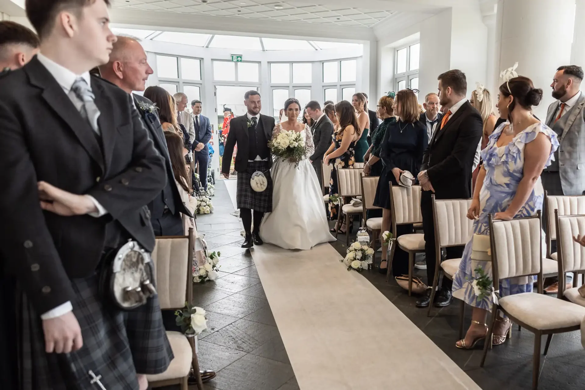 Bride and groom in traditional Scottish attire walk down the aisle, surrounded by guests standing on either side in a decorated indoor venue.