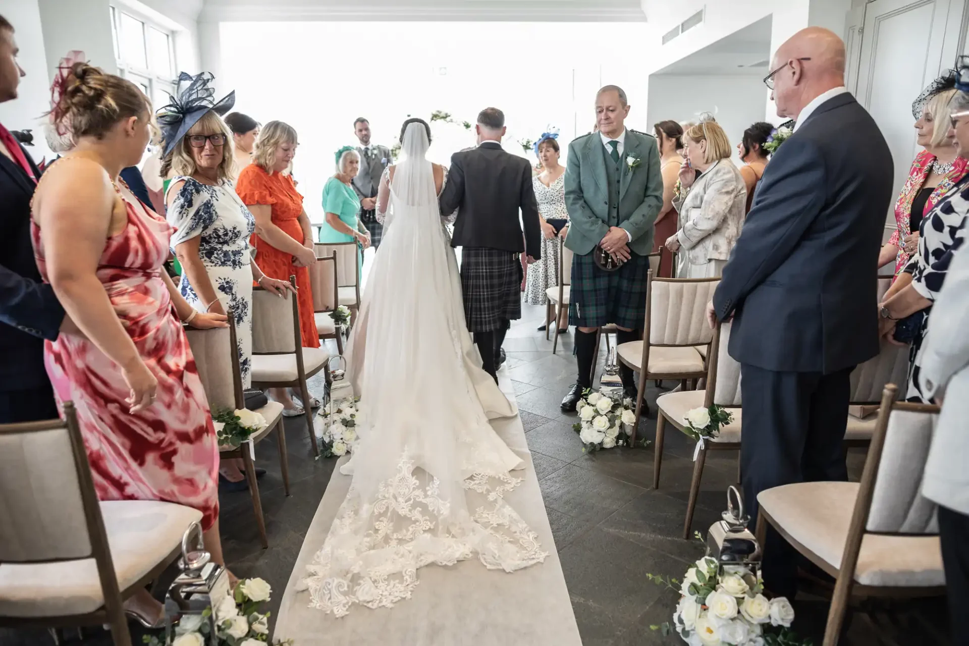 A bride walks down the aisle with a man in a kilt, surrounded by seated guests at a wedding ceremony.