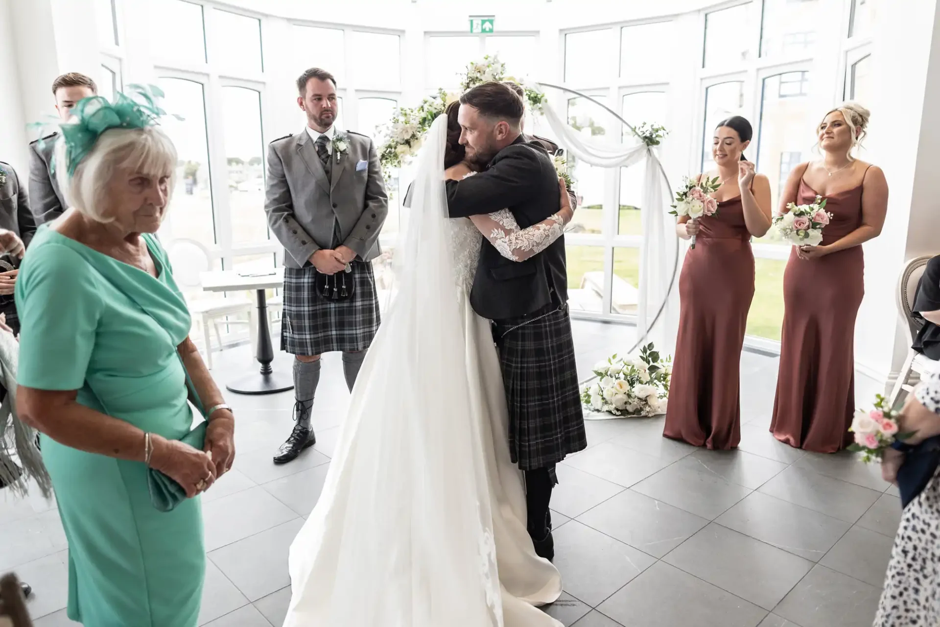 A bride and groom hug in a bright room with bridesmaids and guests nearby. The groom and one guest are wearing kilts. White flowers and natural light fill the background.