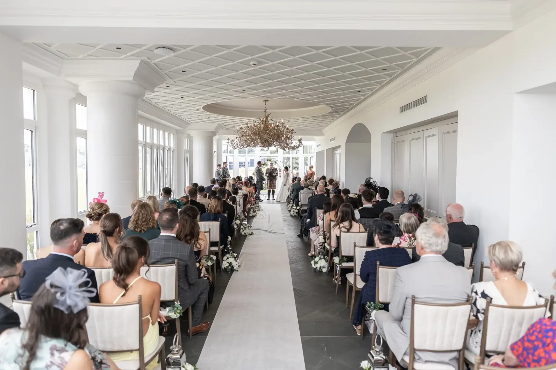 Indoor wedding ceremony with guests seated facing the couple and officiant at the front. The aisle is lined with flowers, and a chandelier hangs from the ceiling.