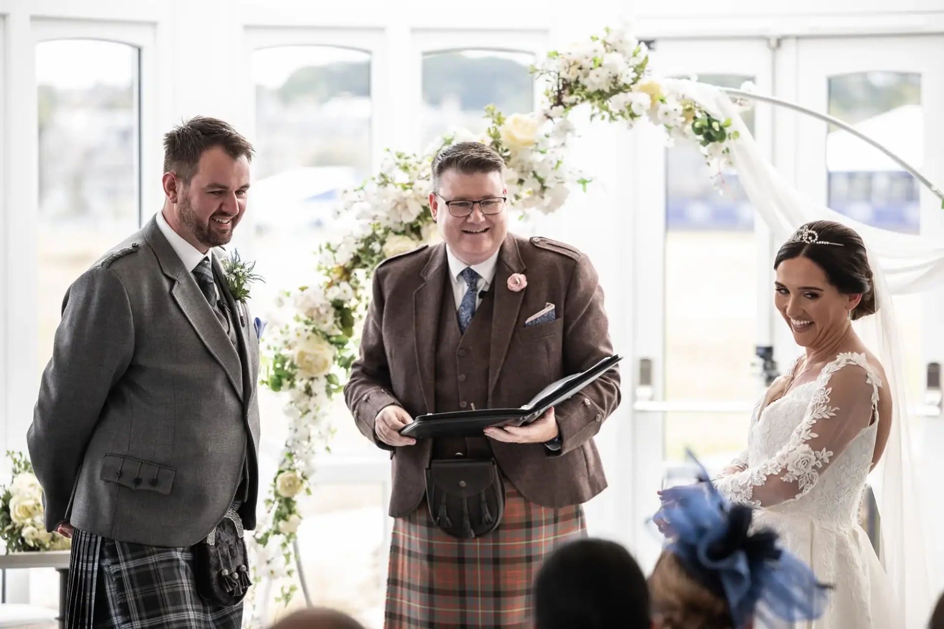 A couple stands with a celebrant in front of a floral arch during a wedding ceremony.