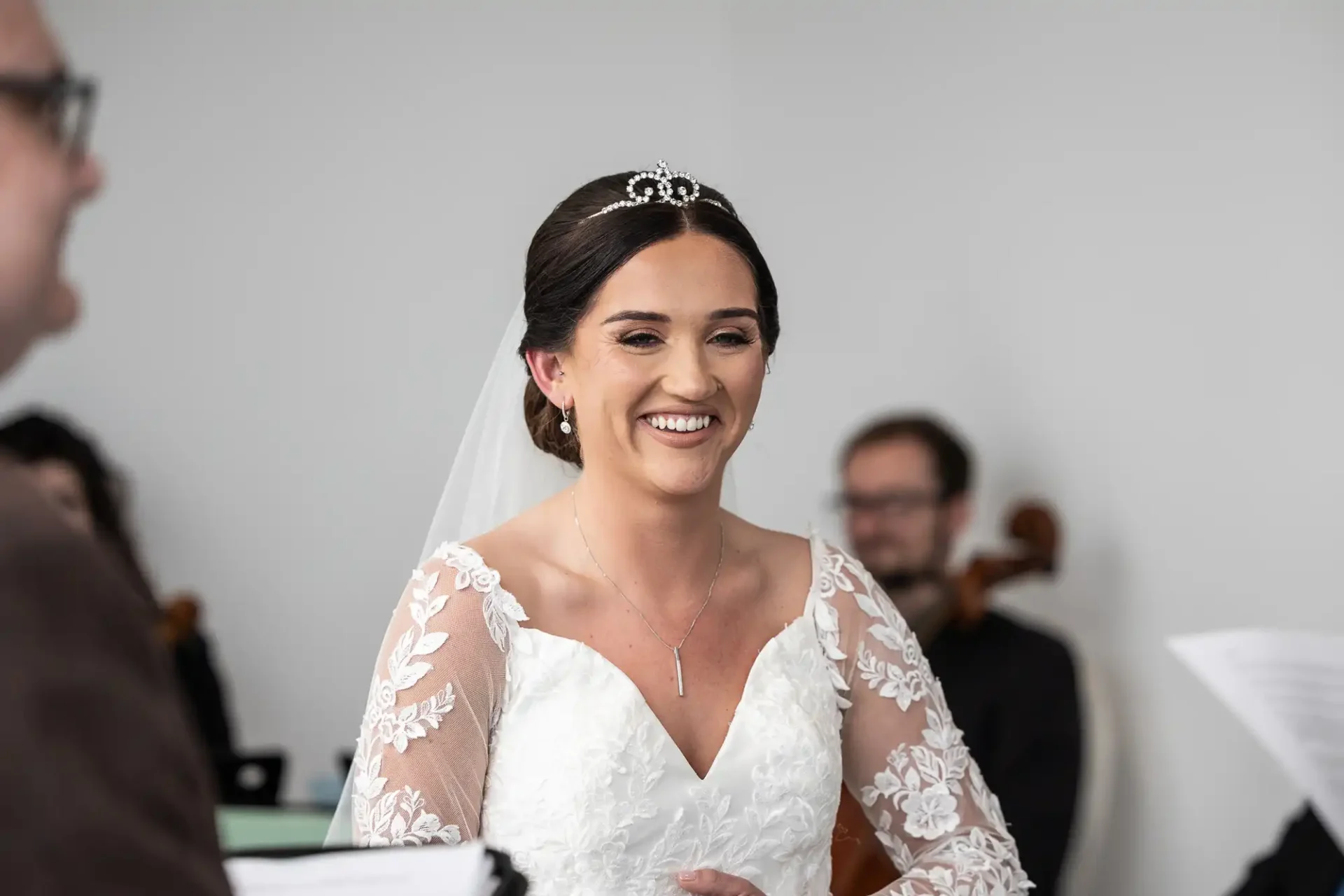 A bride in a white dress and tiara smiles during a ceremony. Musicians with string instruments are visible in the background.