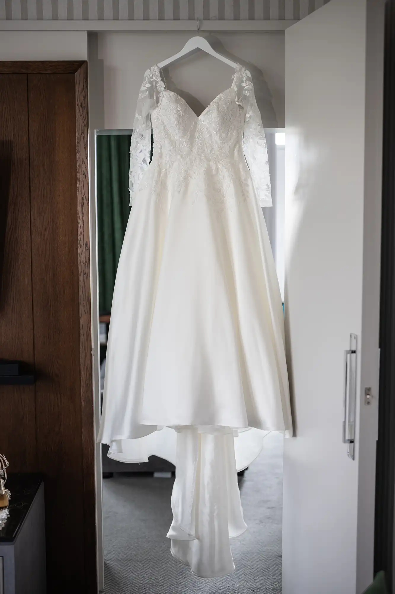 White wedding dress with lace long sleeves and a fitted bodice, hanging on a hanger in a room against a door.