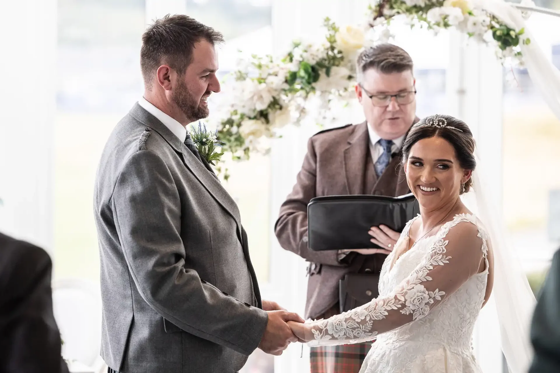 A couple holding hands during a wedding ceremony, with an officiant in the background. The setting is decorated with white flowers.