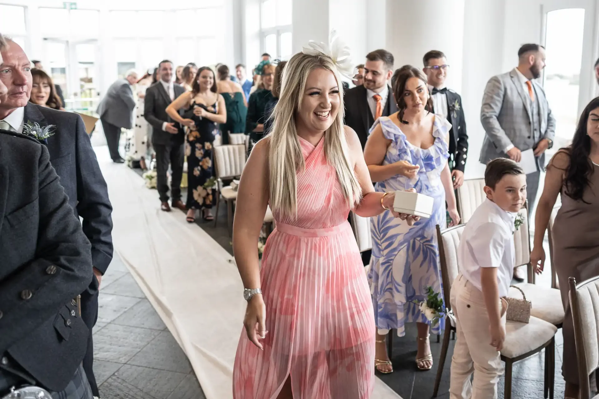 Woman in a pink dress smiles while walking down an aisle at a gathering with seated and standing attendees.