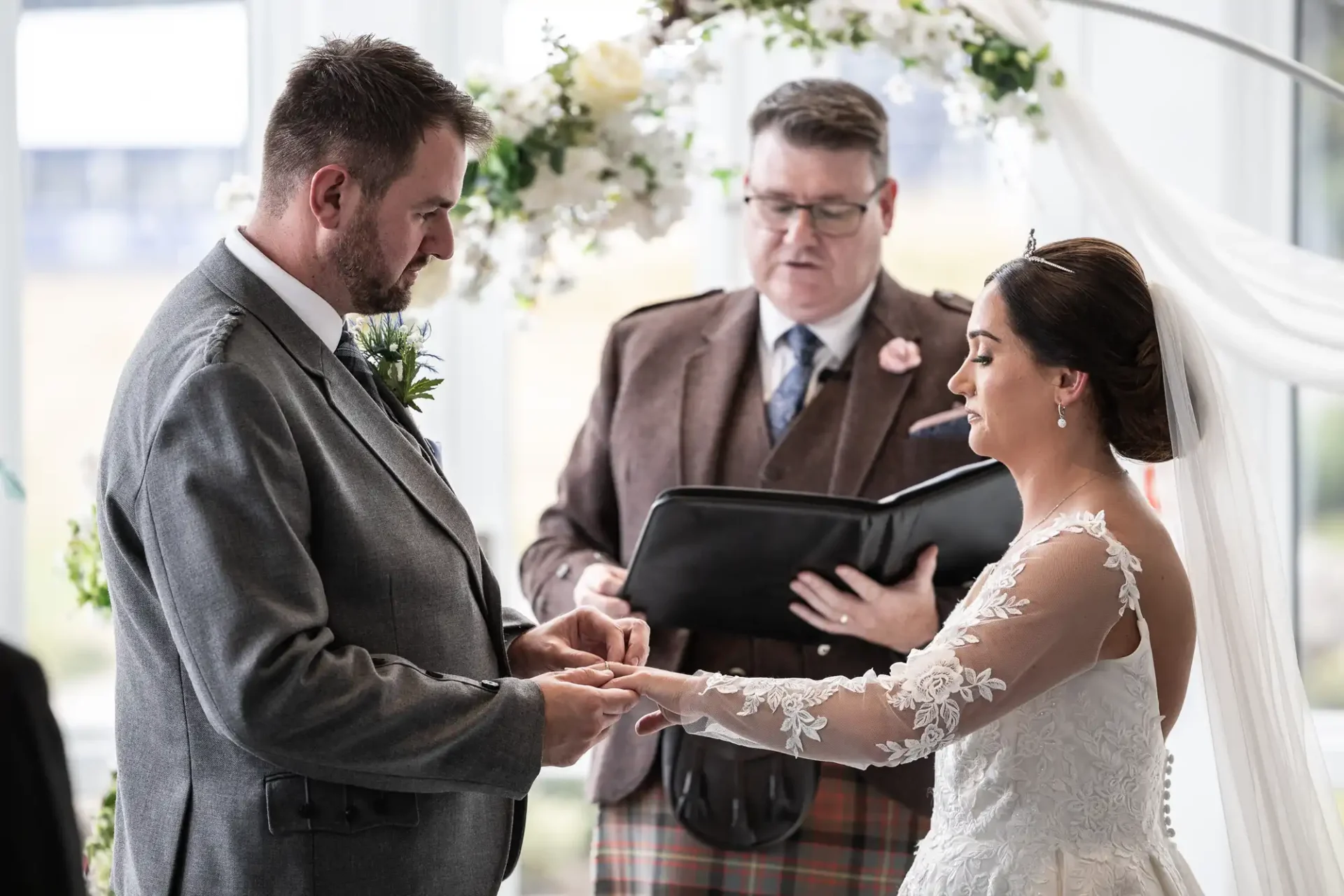 A couple exchanges rings during a wedding ceremony, officiated by a man in a suit. The woman wears a white dress, and the man wears a gray suit. Floral decorations are visible in the background.