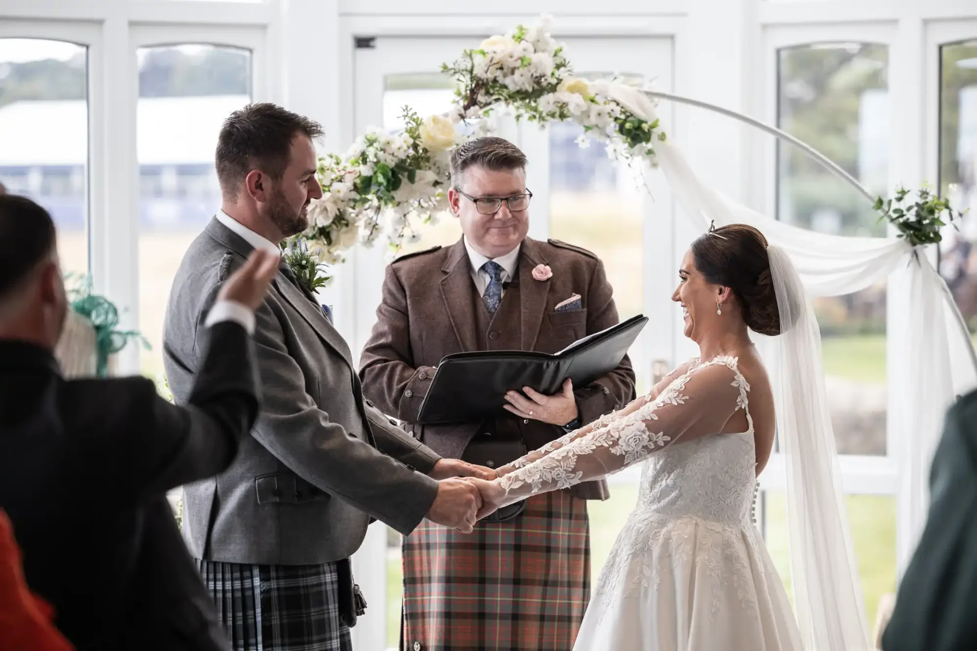 A couple holds hands during a wedding ceremony, officiated by a man in a plaid kilt. They stand under a floral arch indoors, with guests in attendance.