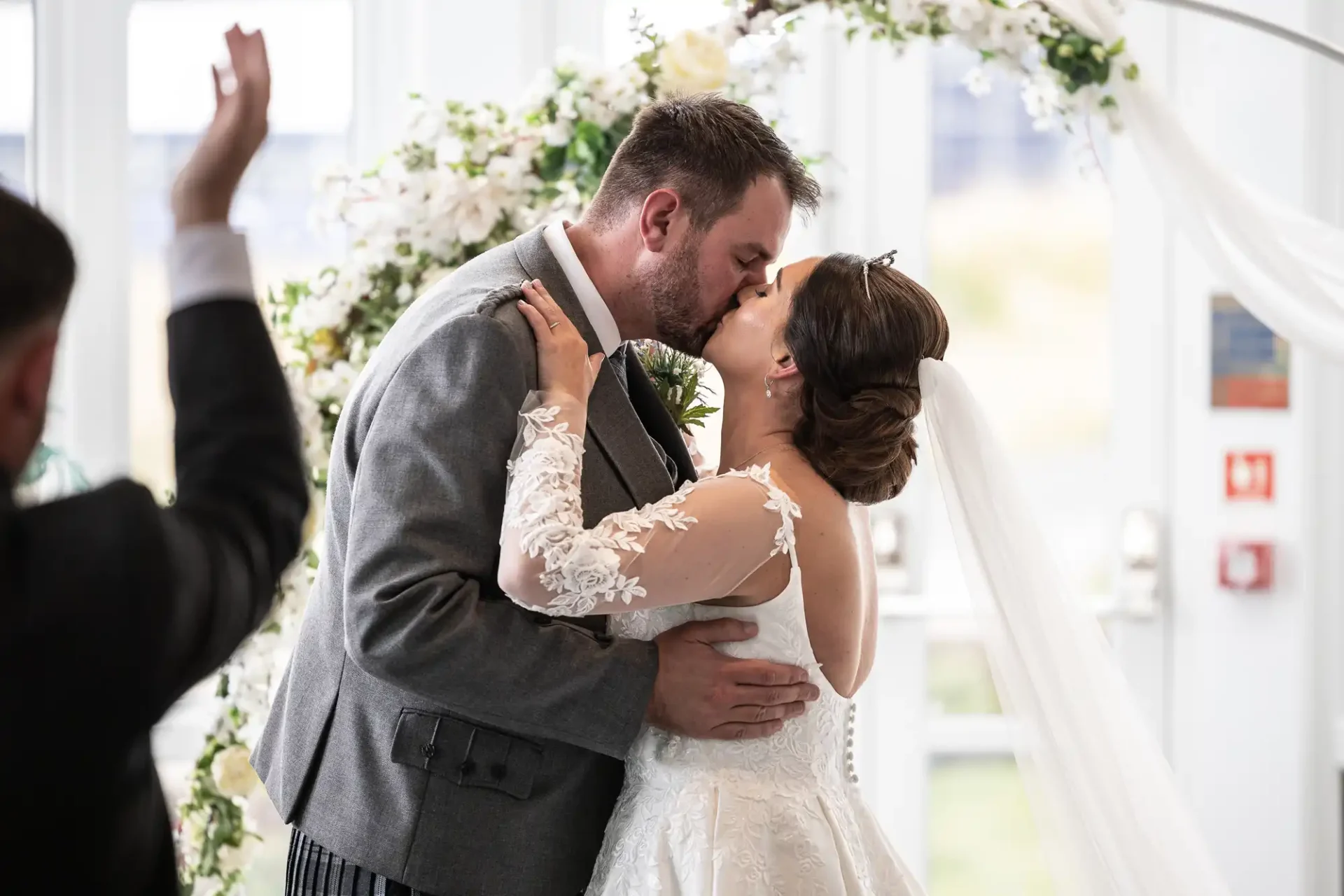 A couple kisses at their wedding ceremony, standing before a floral arch. The bride wears a white gown, and the groom is in a grey suit. A person raises a hand in the background.