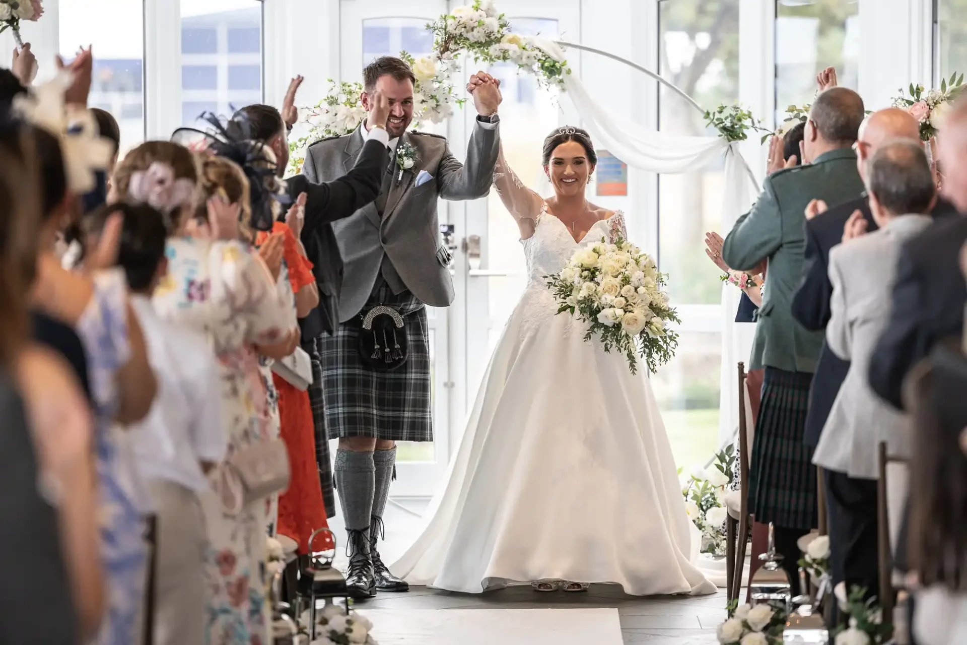 A bride and groom smile and wave while walking down the aisle, surrounded by cheering guests. The groom is wearing a kilt, and the bride is holding a bouquet of white flowers.