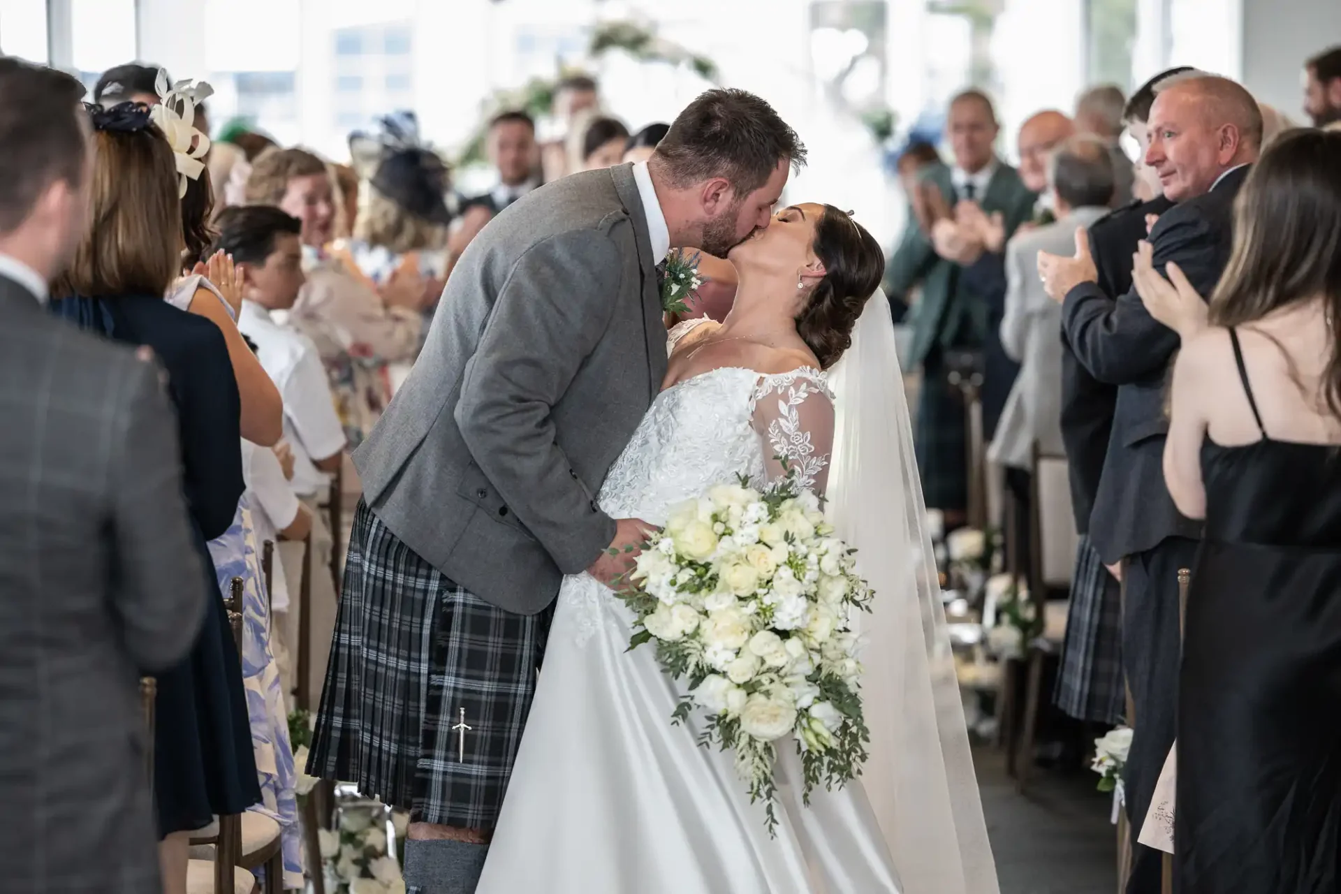 A couple kisses at their wedding ceremony. The groom wears a gray jacket and kilt; the bride wears a white dress and veil. Guests are applauding in the background.