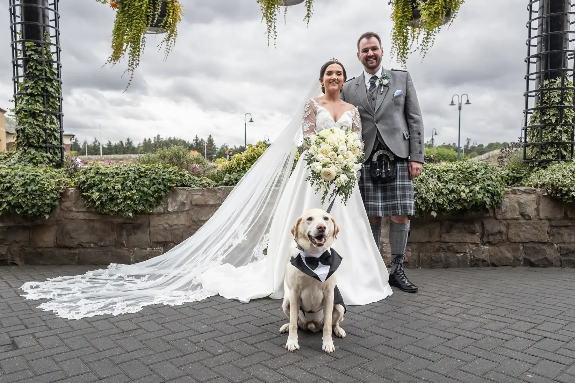 Bride and groom pose outdoors with a dog in a bow tie. Bride wears a white dress and holds a bouquet; groom wears a kilt and grey jacket. Overcast sky and greenery in the background.