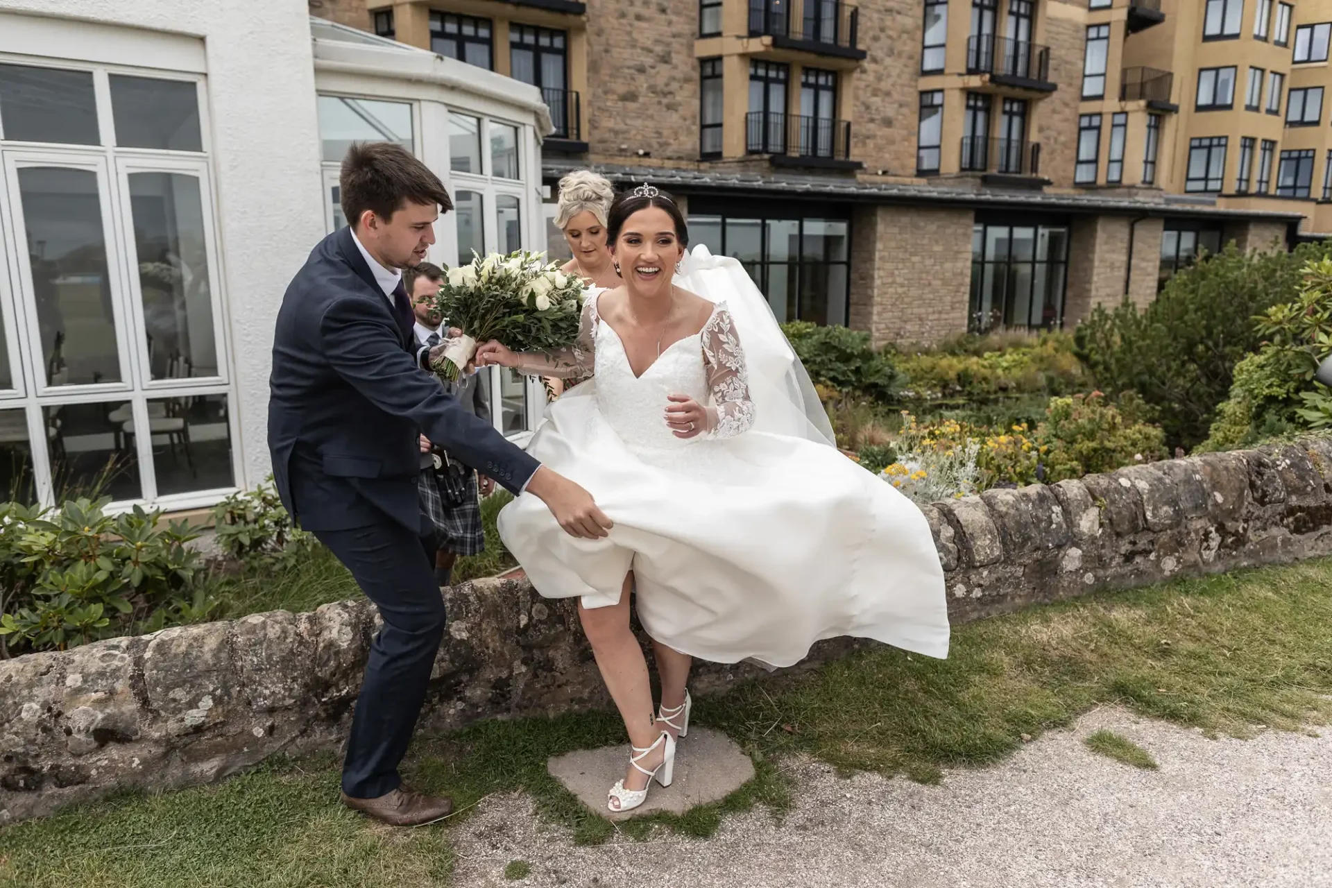 Bride being assisted over a stone wall by two people, with a hotel in the background. She smiles while holding her dress.