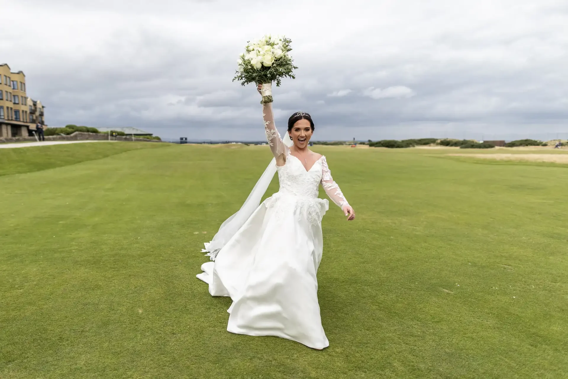 A bride in a white dress raises her bouquet while walking on a green lawn under a cloudy sky.