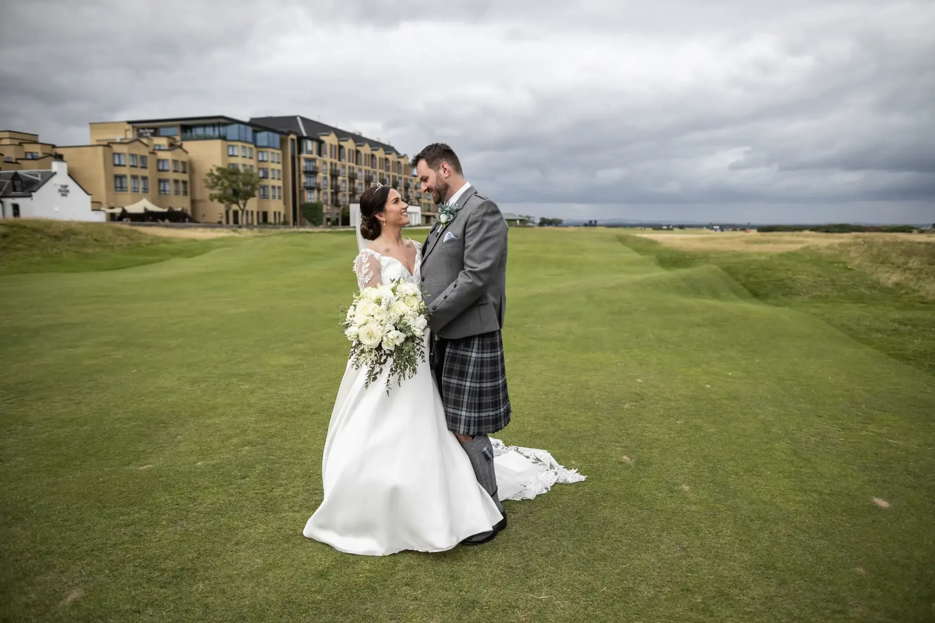 A bride and groom stand on a grassy field, with a building and cloudy sky in the background. The bride holds a bouquet of white flowers, and the groom wears a suit with a tartan kilt.