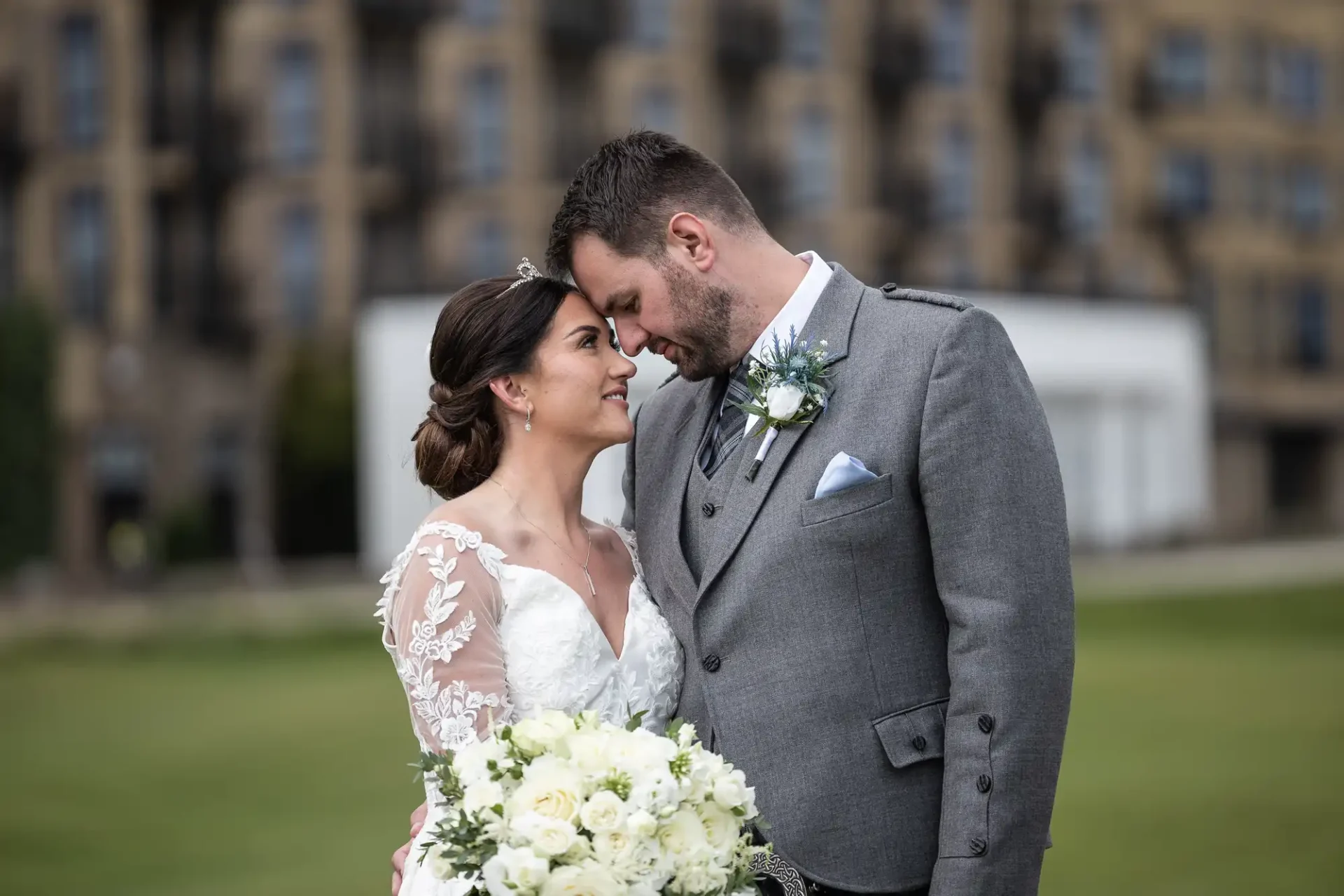 Bride and groom stand closely together outdoors, with the groom in a gray suit and the bride in a white lace dress holding a bouquet of white flowers.