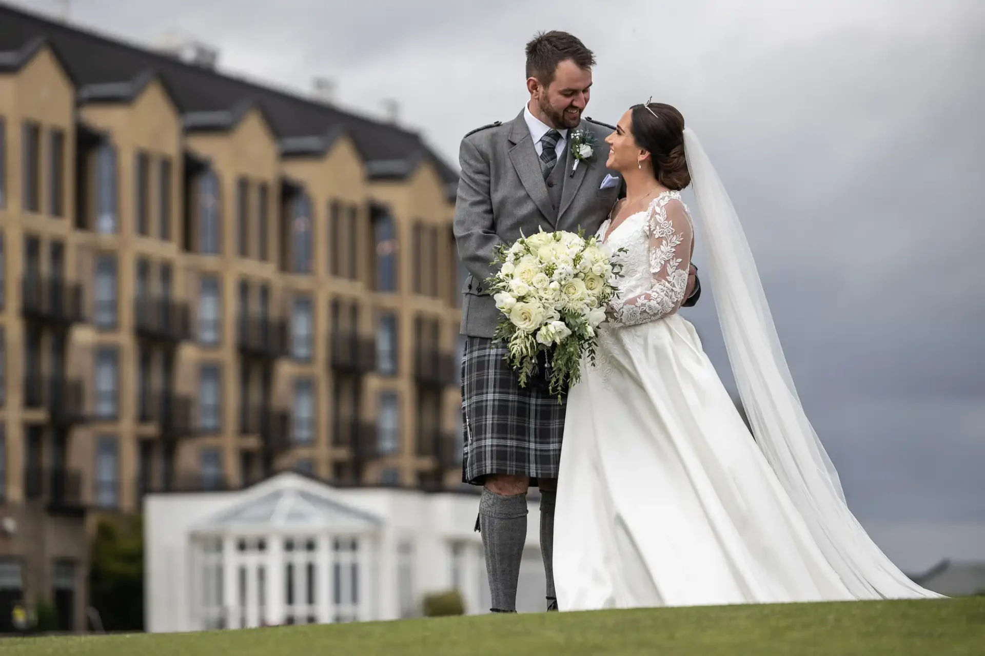 A bride and groom stand on a grassy area, smiling at each other. The groom is in a kilt and jacket, and the bride is in a white dress holding a bouquet. A building is in the background.