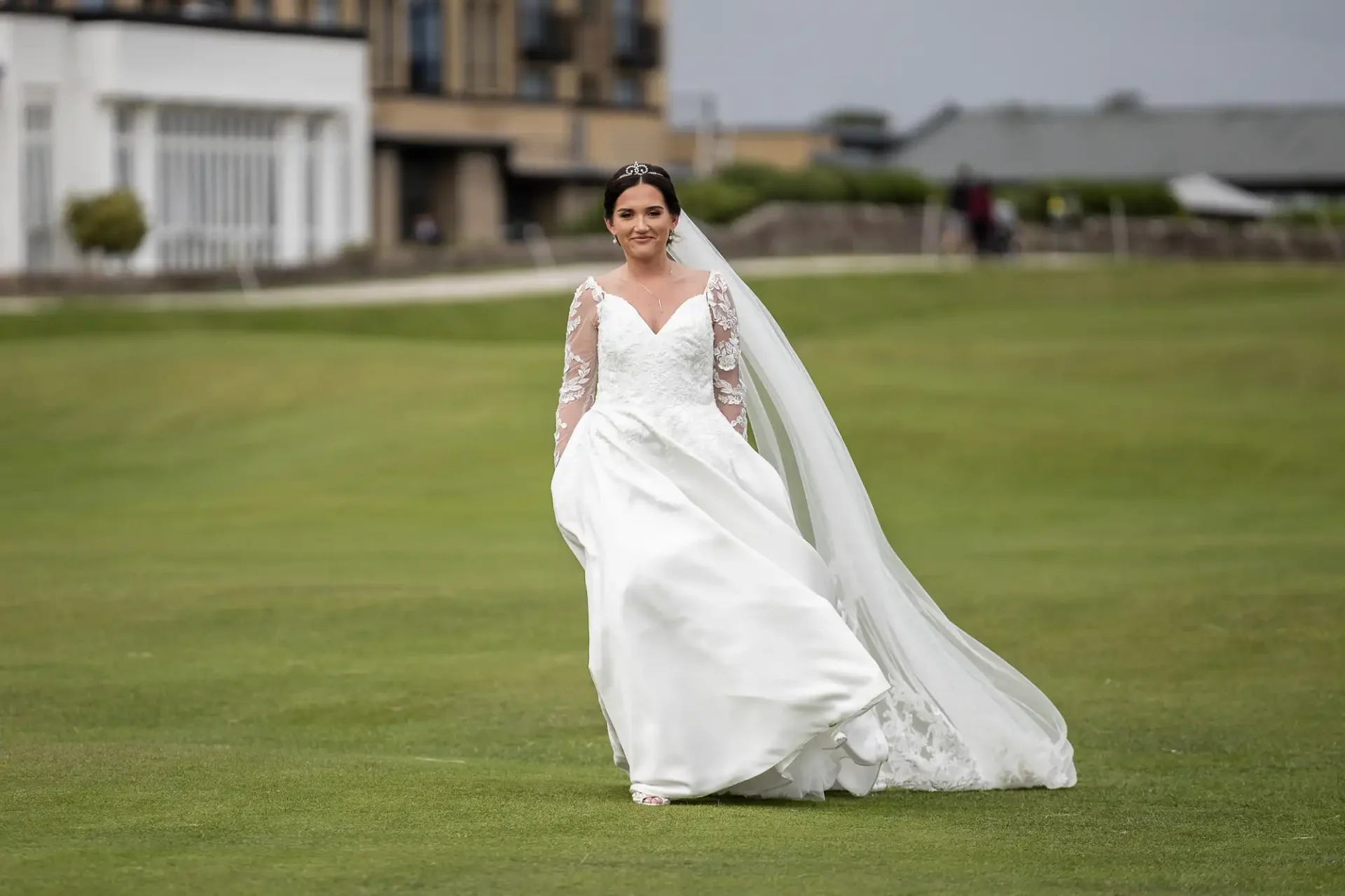 Bride in a white gown and veil walking on grass in front of buildings, smiling.