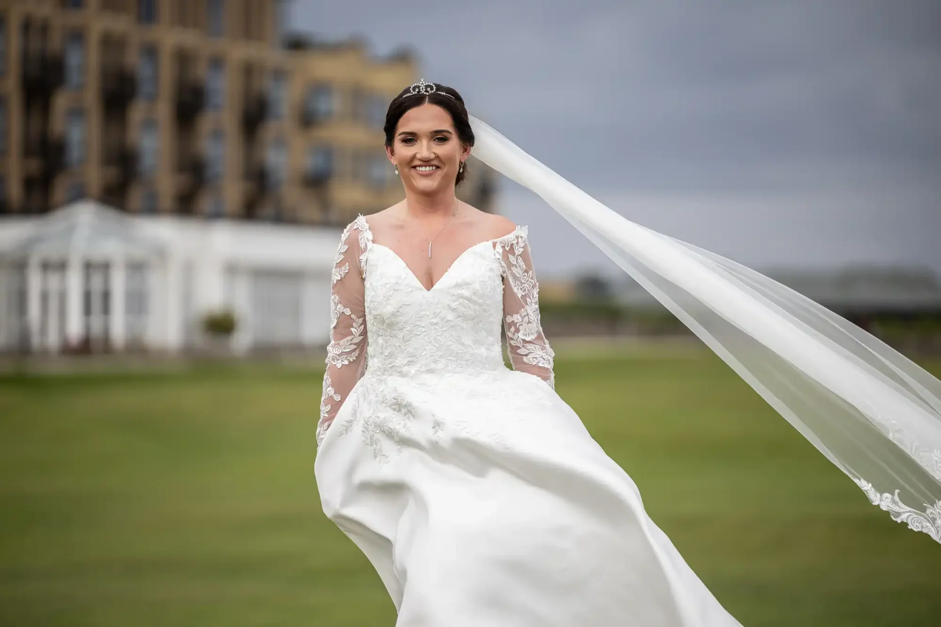 A bride in a white wedding dress with lace sleeves and a long veil stands on grass with a building in the background.