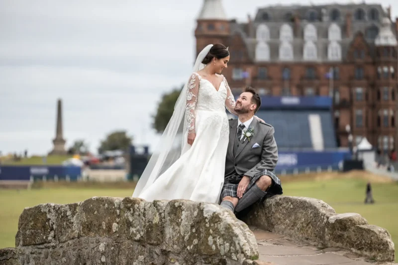 Old Course Hotel wedding photos: Bride in a white gown and groom in a kilt sitting on a stone bridge, with a historic building and obelisk in the background.