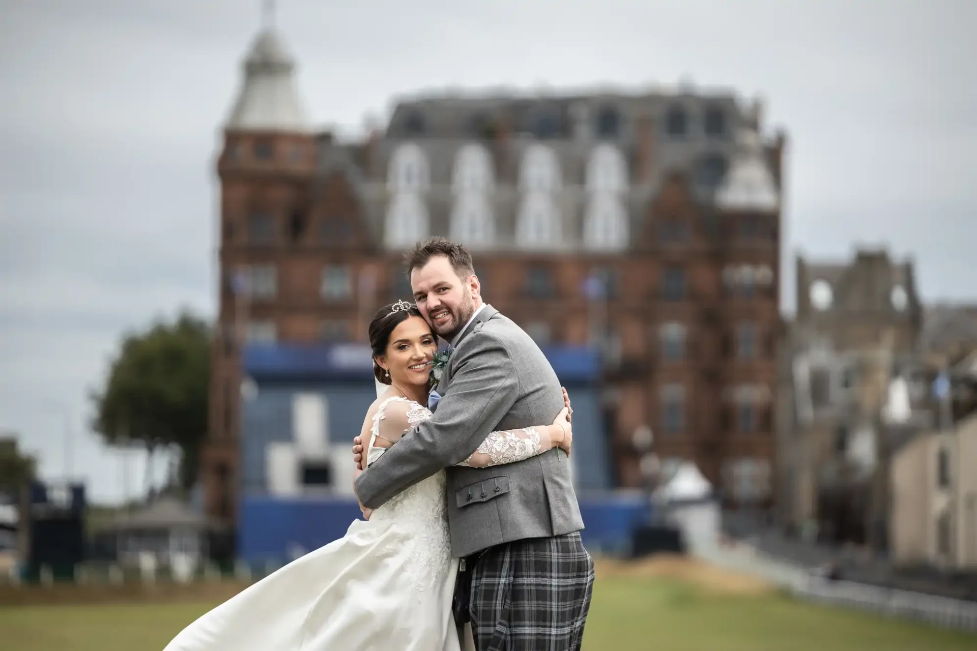 Bride and groom embracing on grass, with an ornate brick building and cloudy sky in the background.