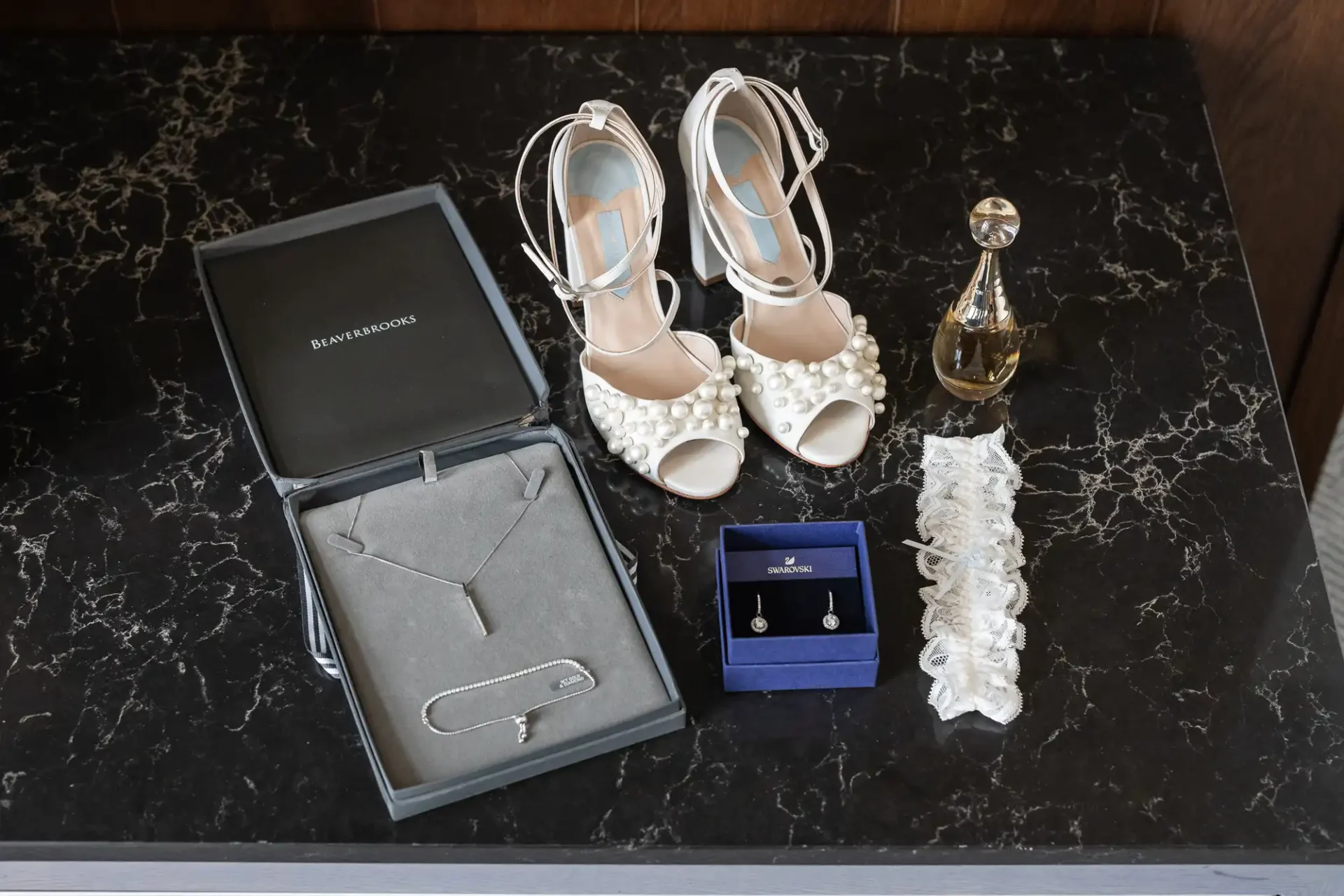 Open shoe box with pearl-embellished heels, a perfume bottle, necklace in case, earrings in a box, and a lace garter on a black marble surface.