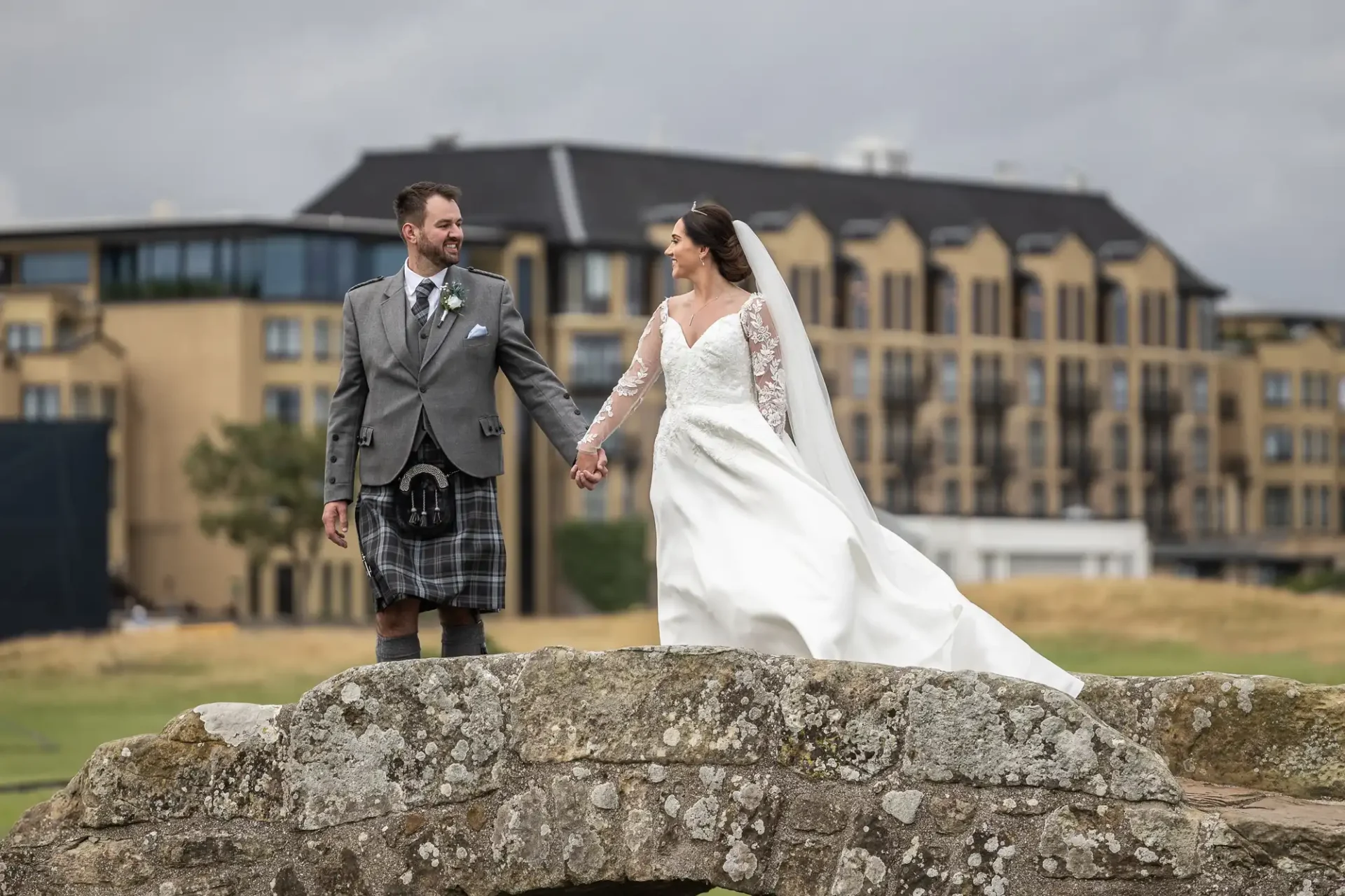 Bride and groom holding hands, standing on a stone bridge. Bride wears a white gown and veil; groom wears a kilt with a grey jacket. Buildings and cloudy sky in the background.