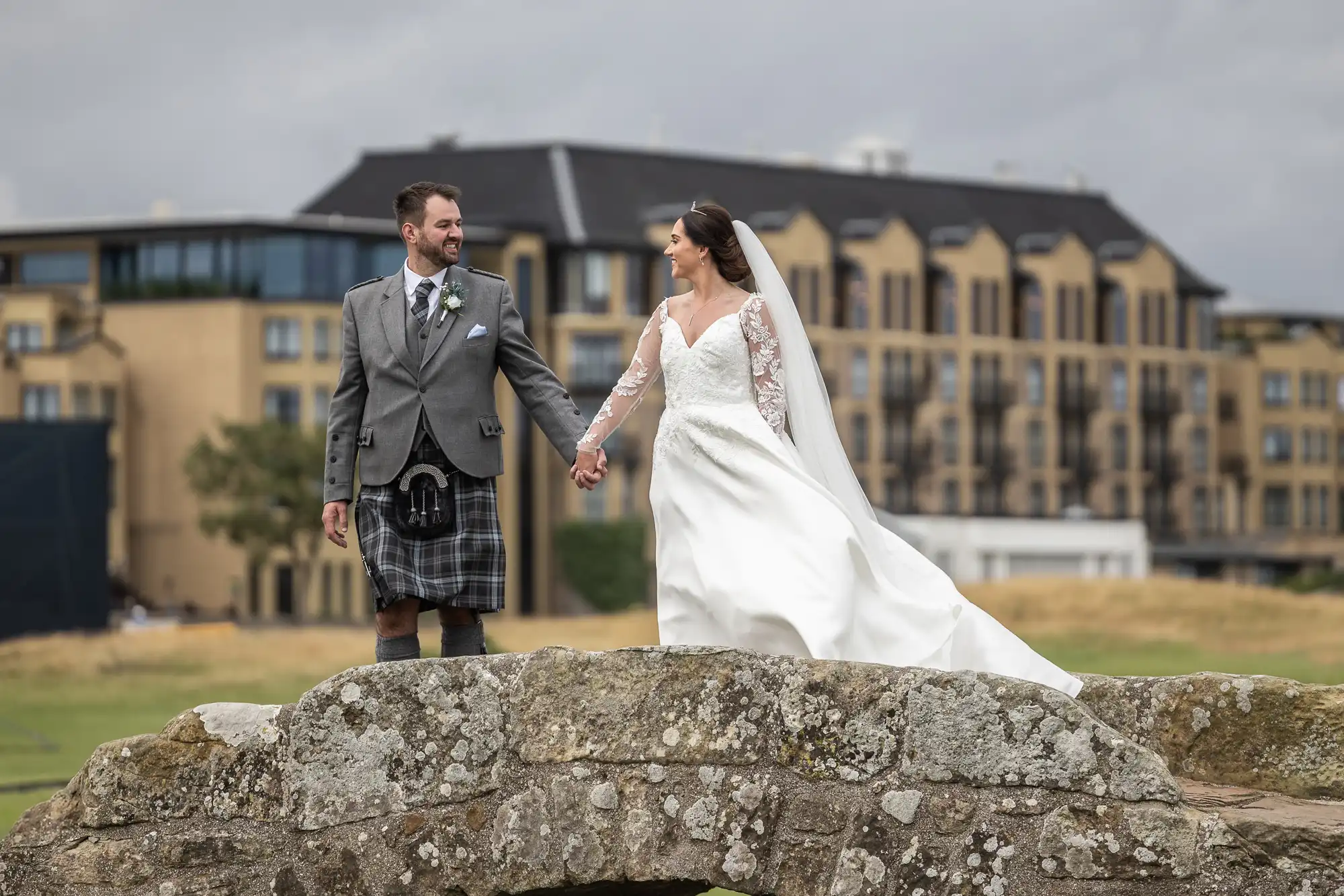 Bride and groom holding hands, standing on Swilken Bridge. Bride wears a white gown and veil; groom wears a kilt with a grey jacket. Buildings and cloudy sky and the Old Course Hotel in the background.