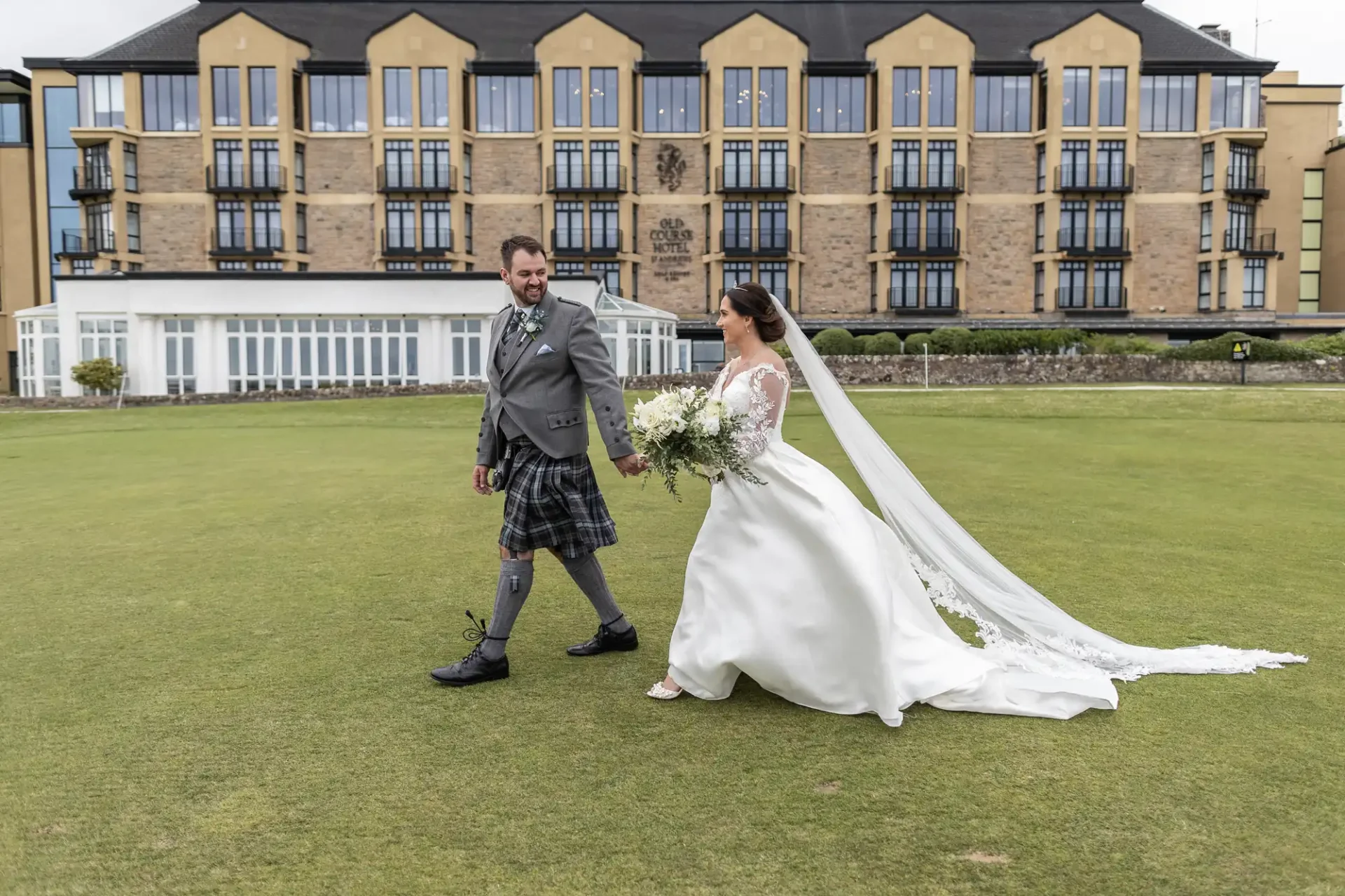 A bride and groom walk hand in hand on grass in front of a large building. The groom wears a kilt, and the bride wears a white gown with a veil, holding a bouquet.