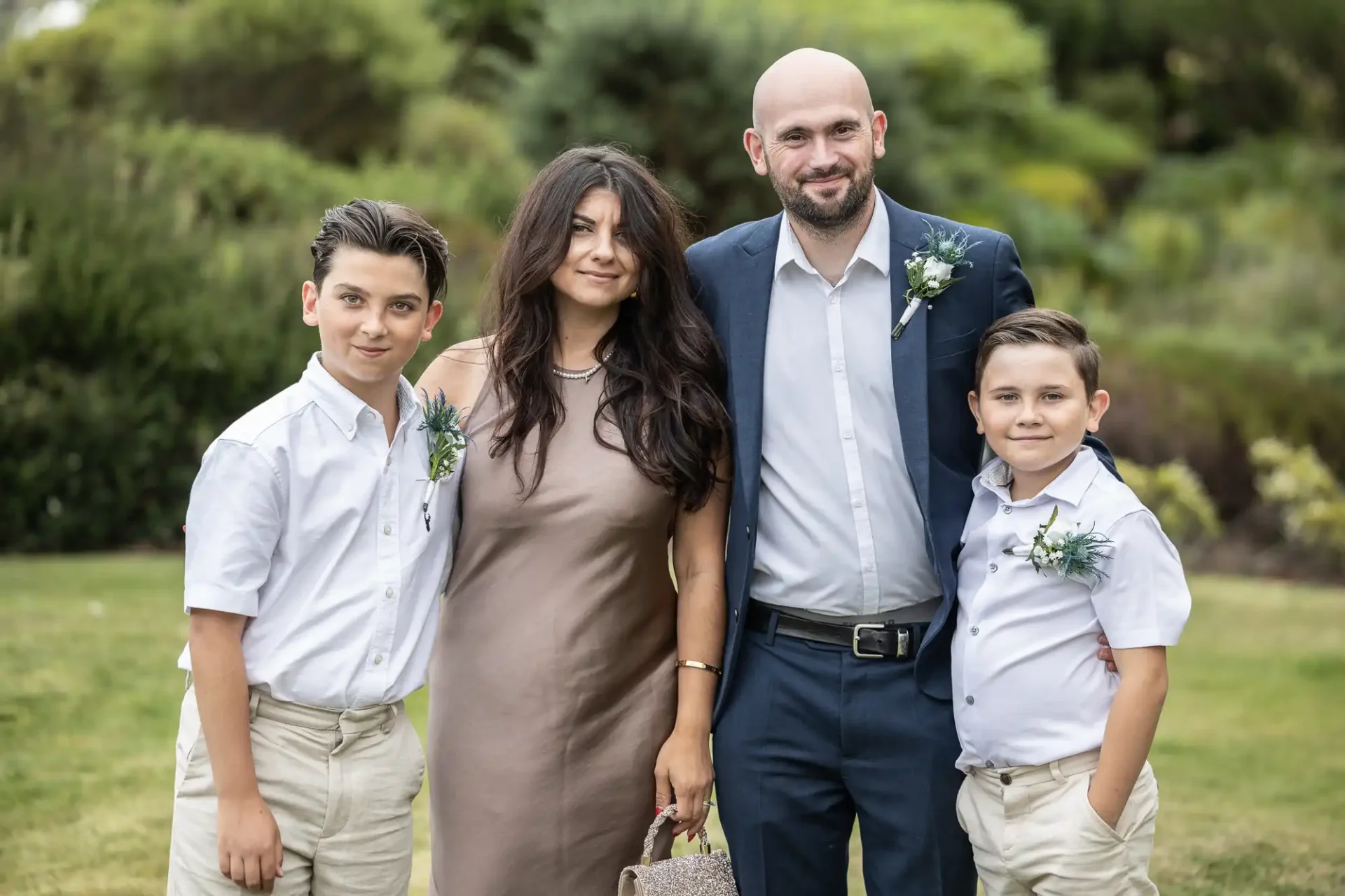 Two adults and two children pose outdoors, dressed in formal attire, with greenery in the background.