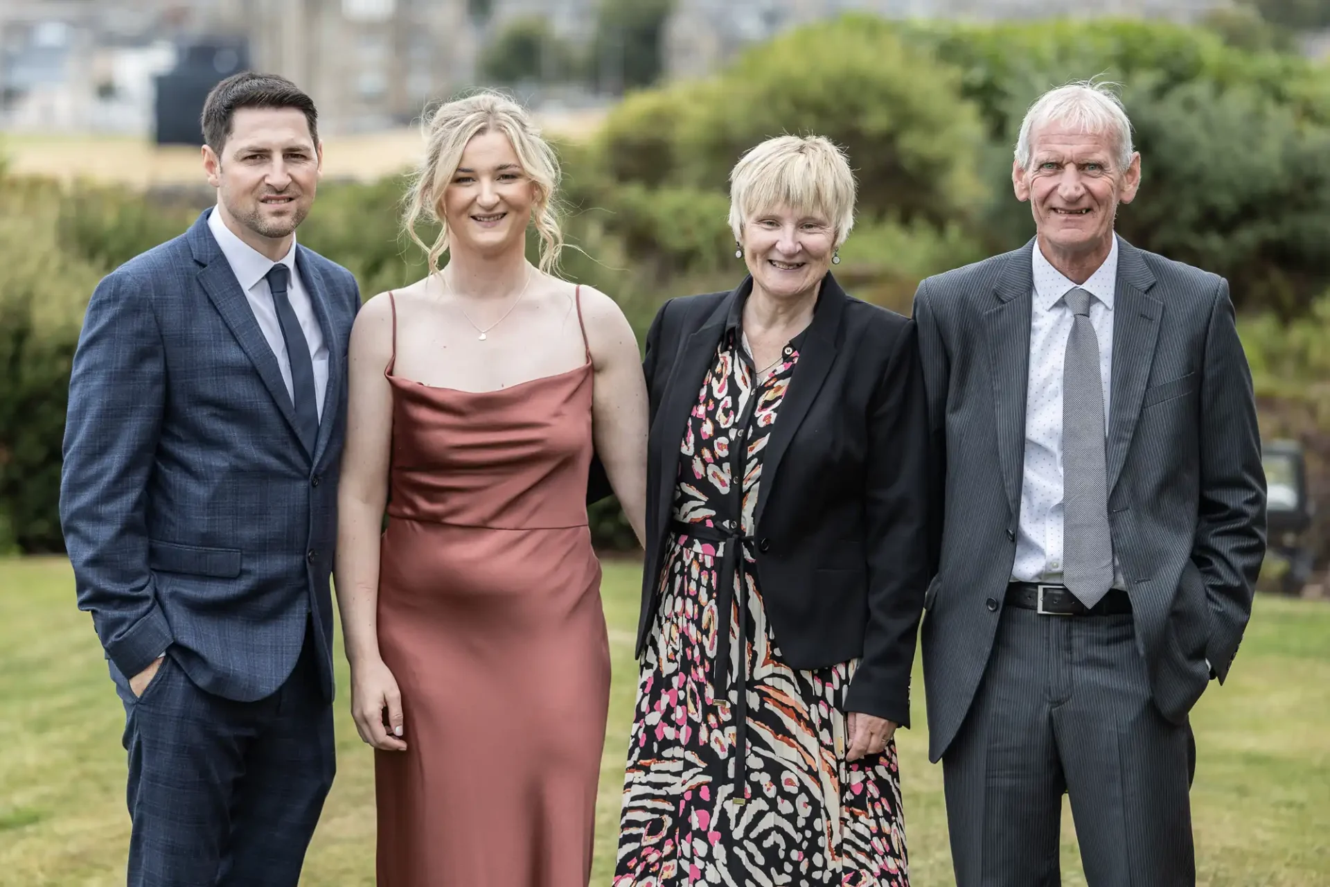 Four people in formal attire stand outside on grass with trees in the background.